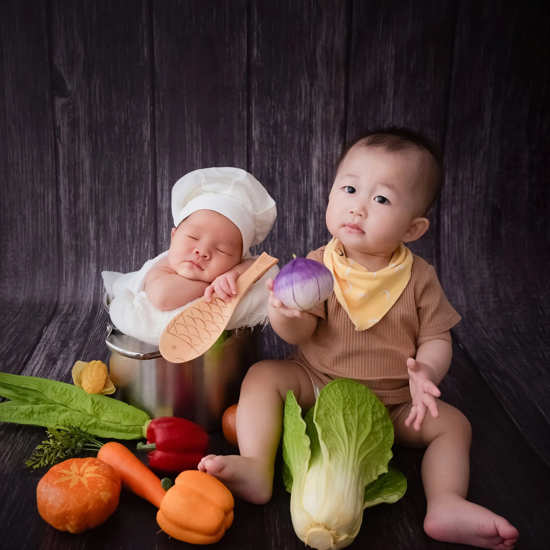 Older sibling holding a purple vegetable prop beside a sleeping newborn in a white chef hat surrounded by colourful vegetables