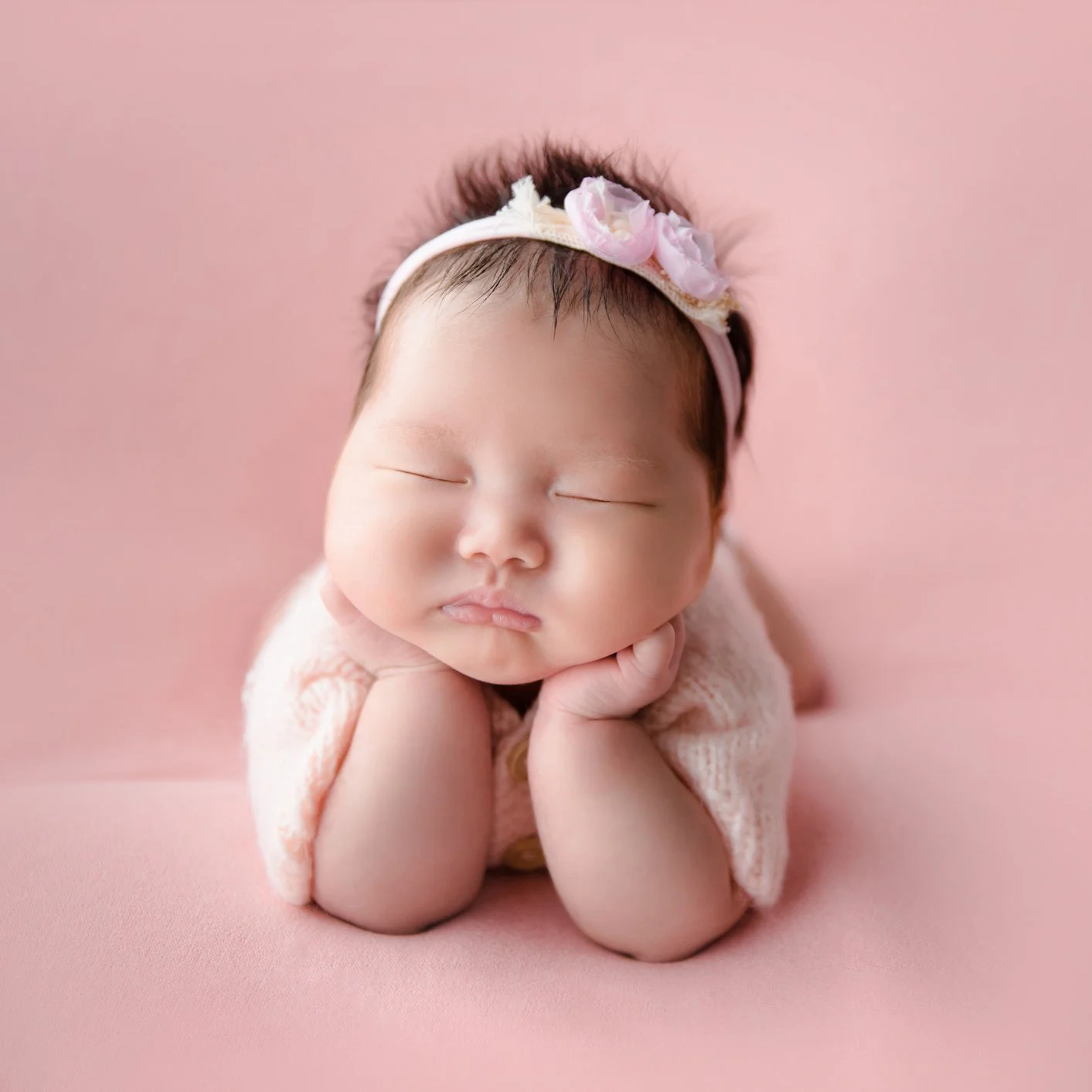 Sleeping newborn wearing a pink flower headband resting both cheeks on hands on a plain pink backdrop