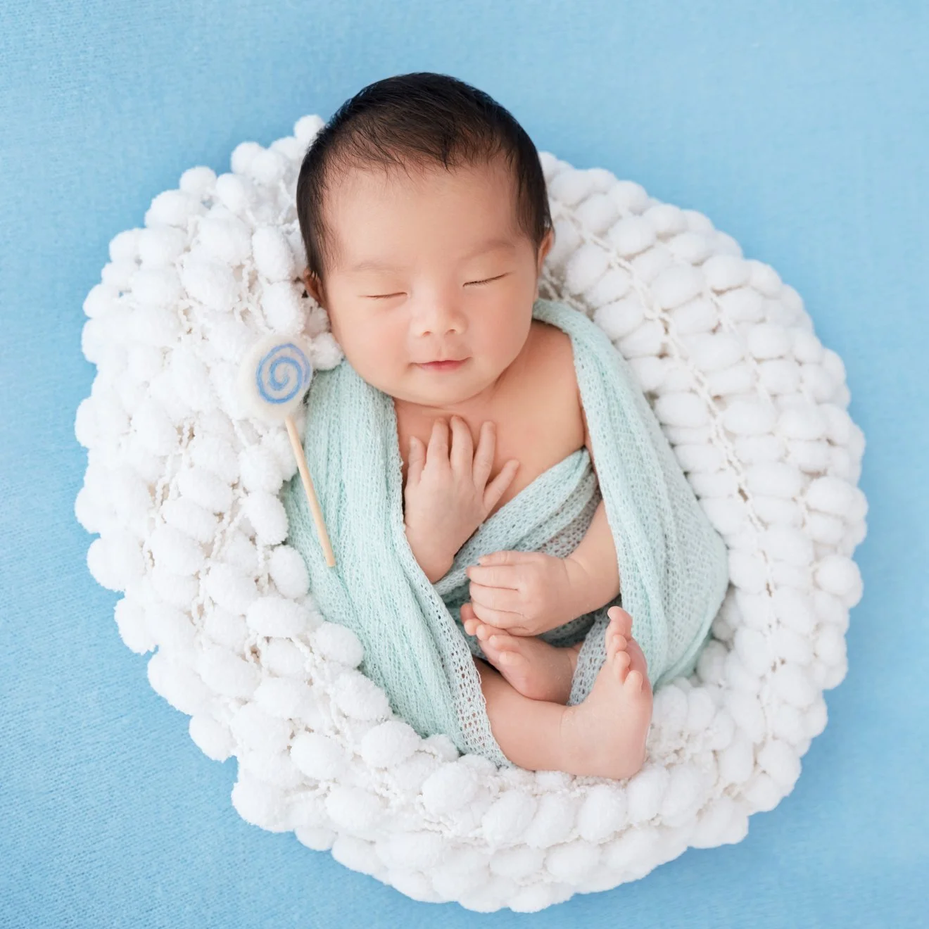 Sleeping newborn in a pale mint knitted wrap reclining with one hand over the chest in a white basket on a blue backdrop
