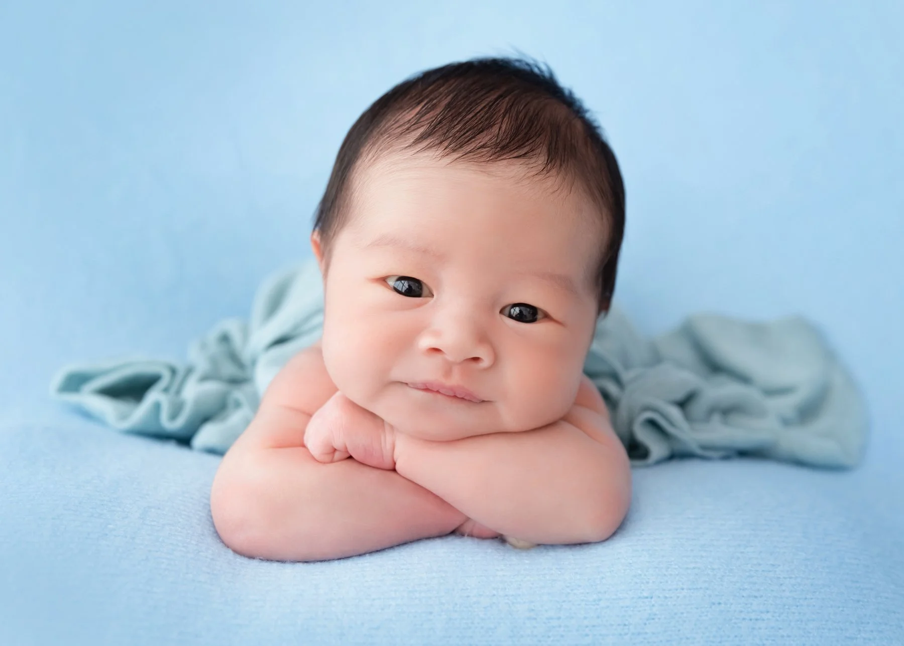 Awake newborn resting the chin on folded arms on a blue blanket with a pale blue wrap behind the body