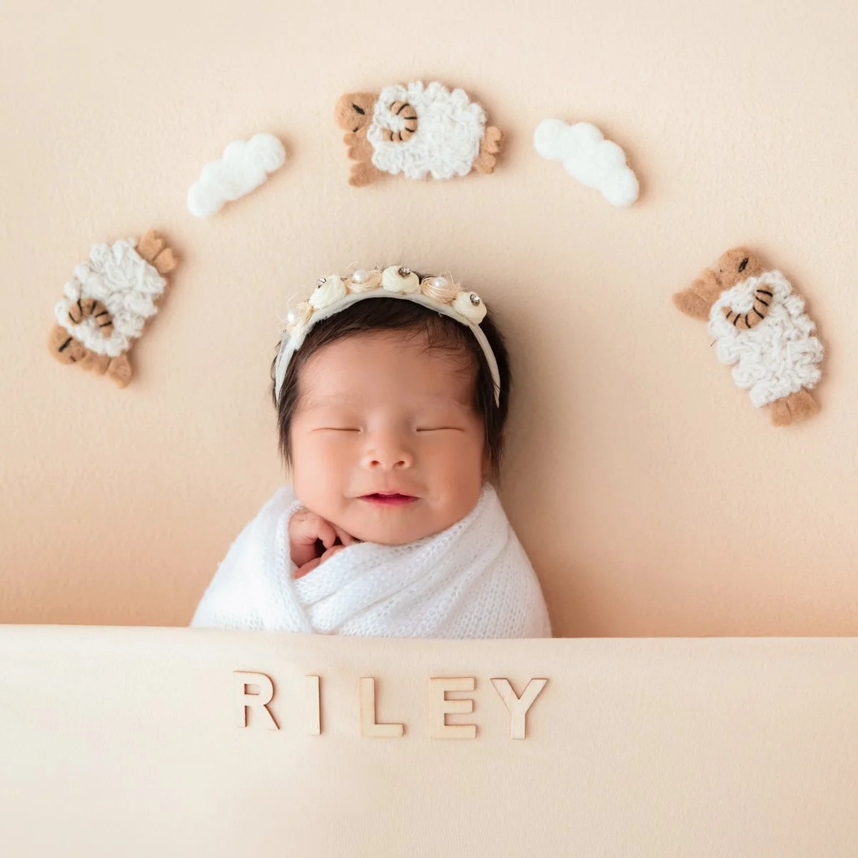 Sleeping newborn in a white swaddle and pearl-flower headband beneath small sheep and cloud decorations on a beige backdrop