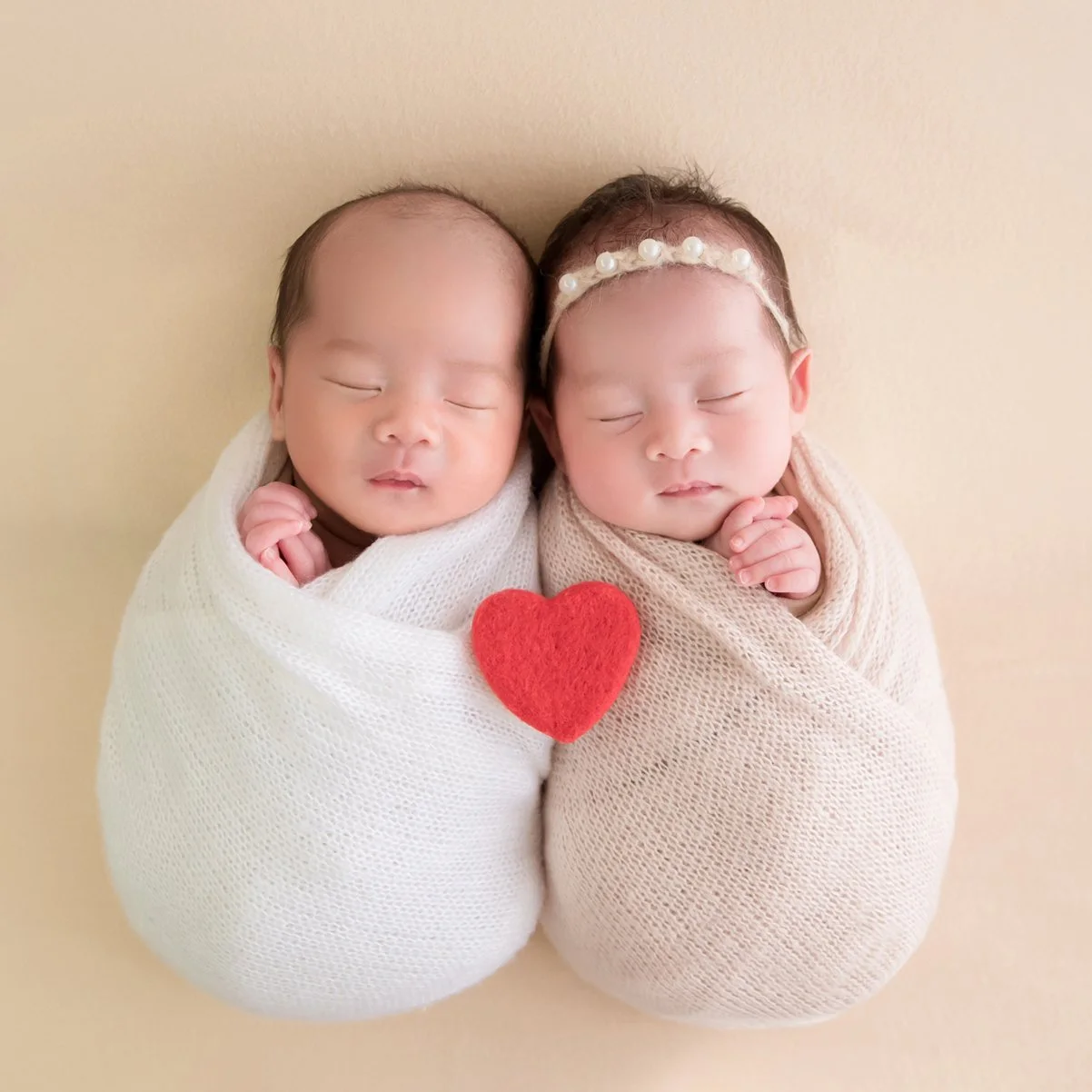 Sleeping newborn twins wrapped in white and beige blankets with a red heart