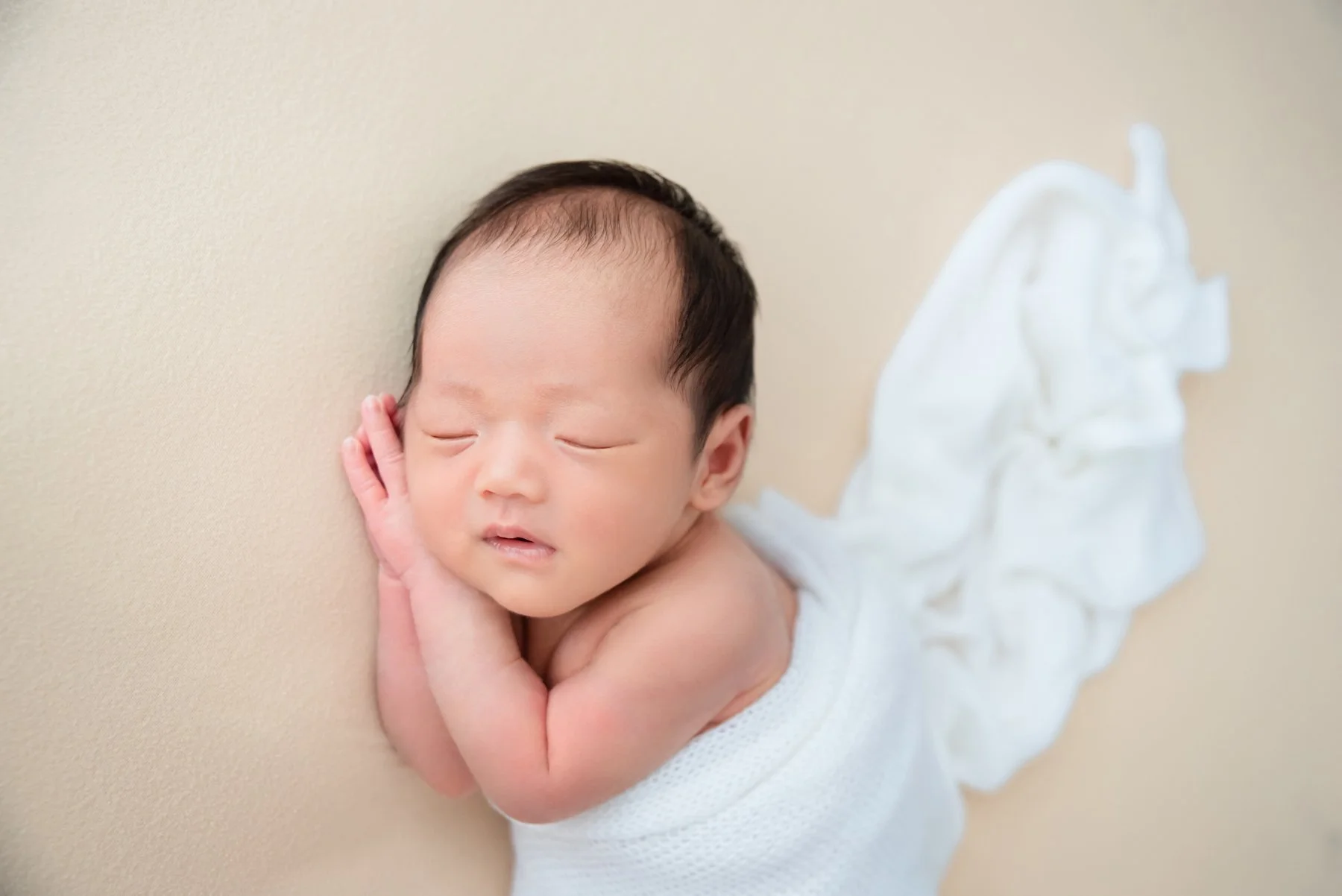 Sleeping newborn in a white wrap lying on one side with hands beside the cheek on a plain beige backdrop