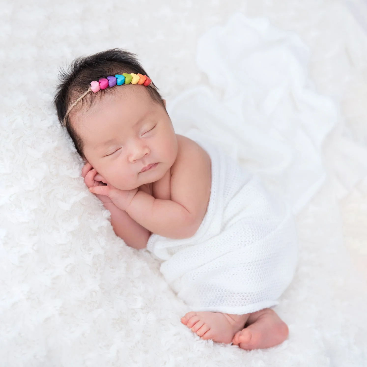 Sleeping newborn in a white wrap and colourful floral headband lying on one side with hands beside the cheek on white fur