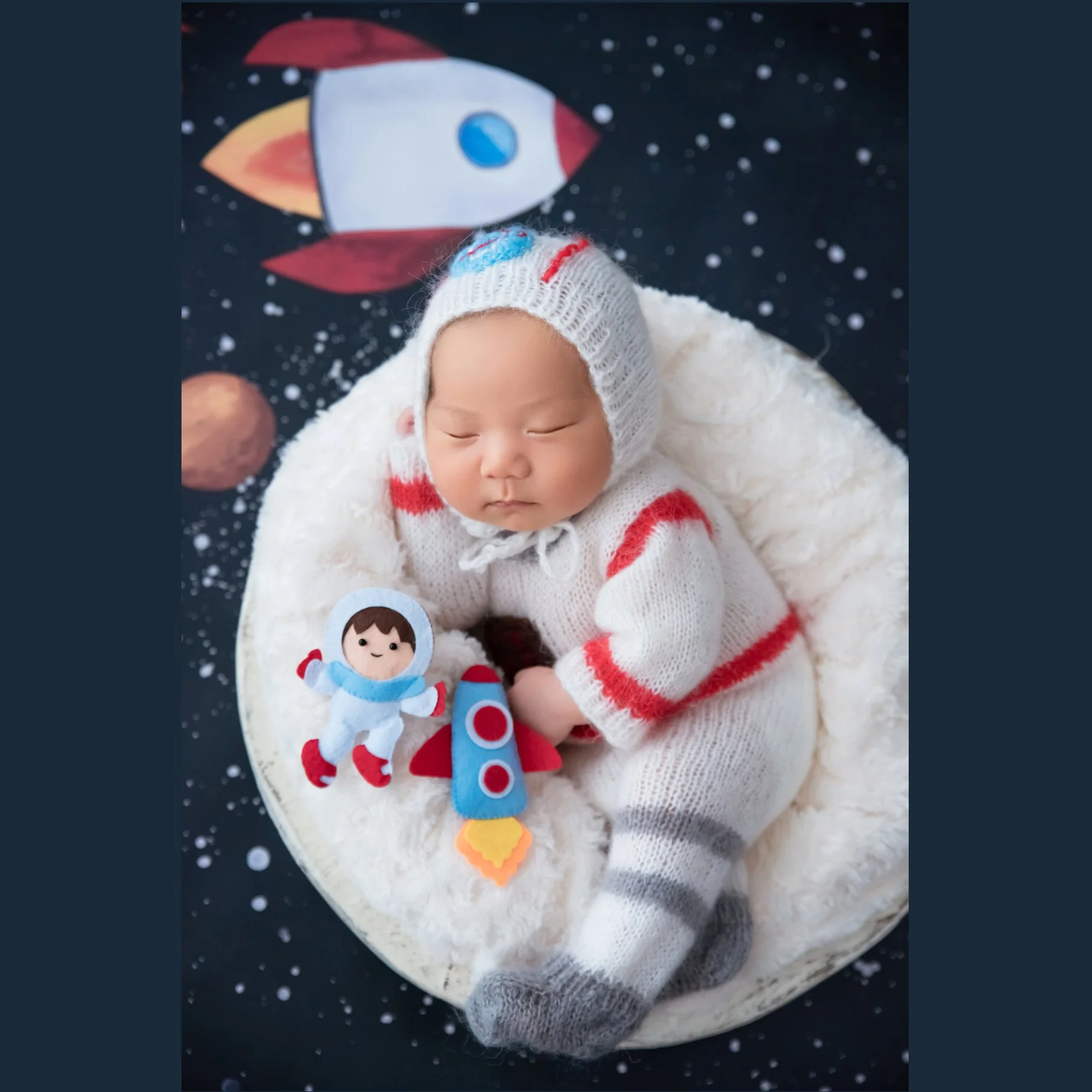 Newborn in a white-and-red astronaut outfit lying on one side beside a toy astronaut and rocket in a fluffy white basket