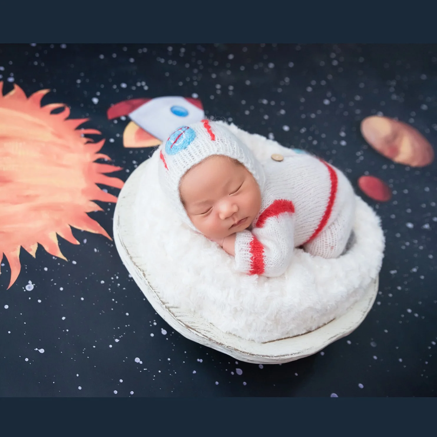 Sleeping newborn in a white astronaut bonnet and outfit resting on folded hands in a fluffy basket on a starry planet-and-rocket backdrop