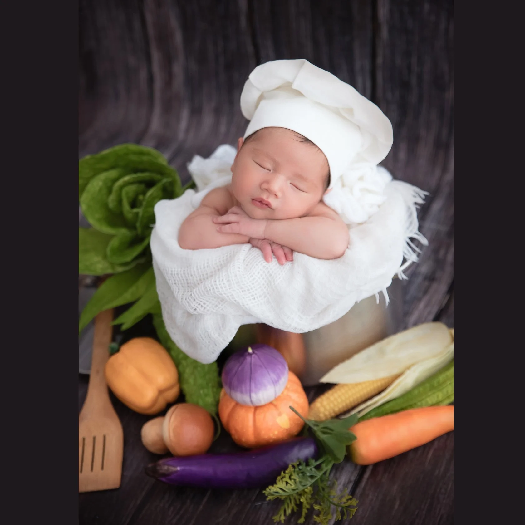 Sleeping newborn in a white chef hat resting on folded arms in a small cooking pot among wooden utensils and vegetable props