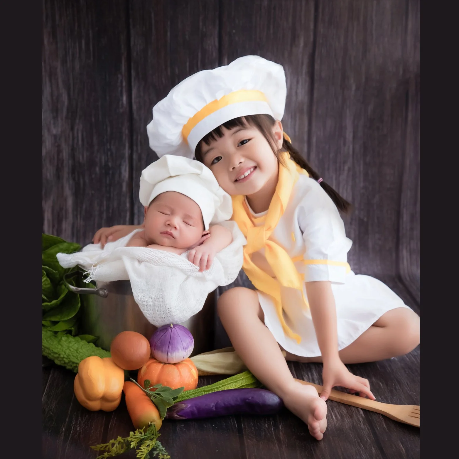 Smiling older sibling in a white chef hat sitting beside a sleeping newborn in a cooking pot with colourful vegetable props