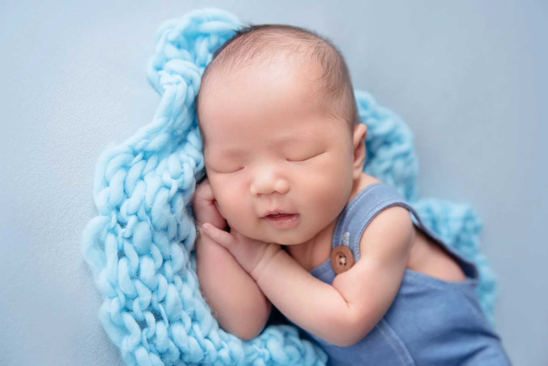 Sleeping newborn with hands beside the cheek nestled in a blue knitted blanket on a pale blue backdrop