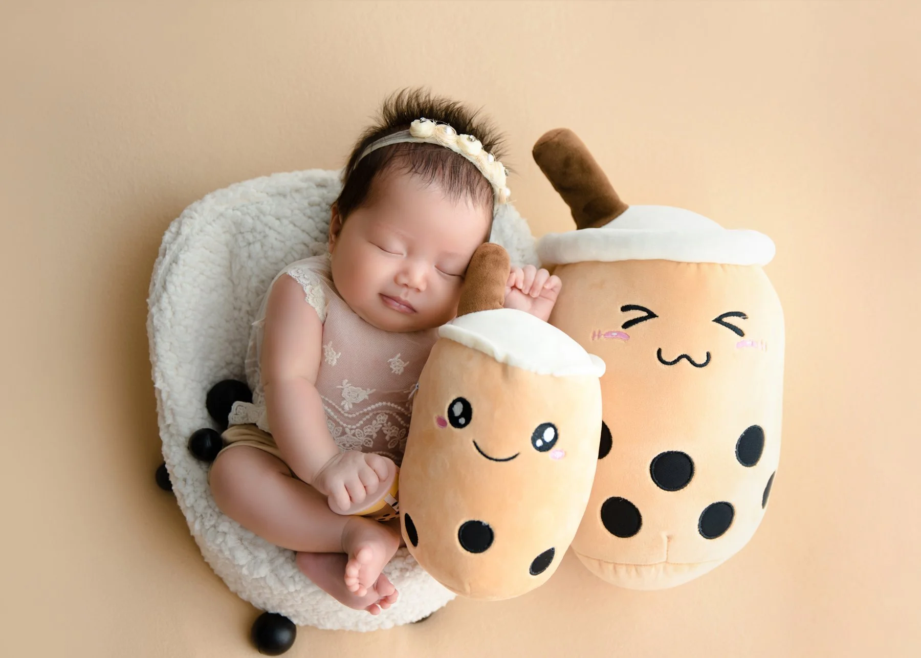 Sleeping newborn wearing a floral headband in a white basket beside two smiling bubble-tea plush cups and black pearls