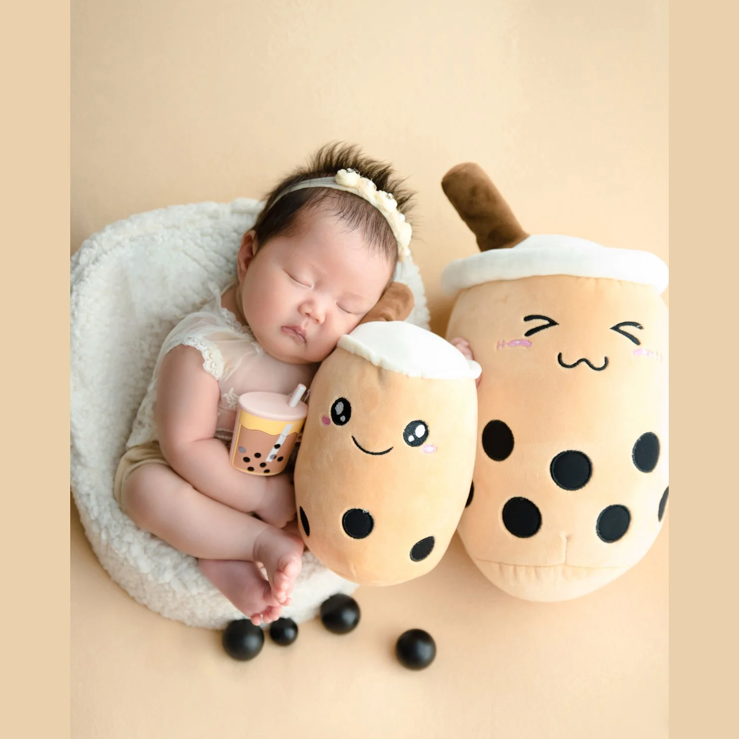 Sleeping newborn with a floral headband cuddling a miniature bubble-tea cup beside two plush cups in a white basket