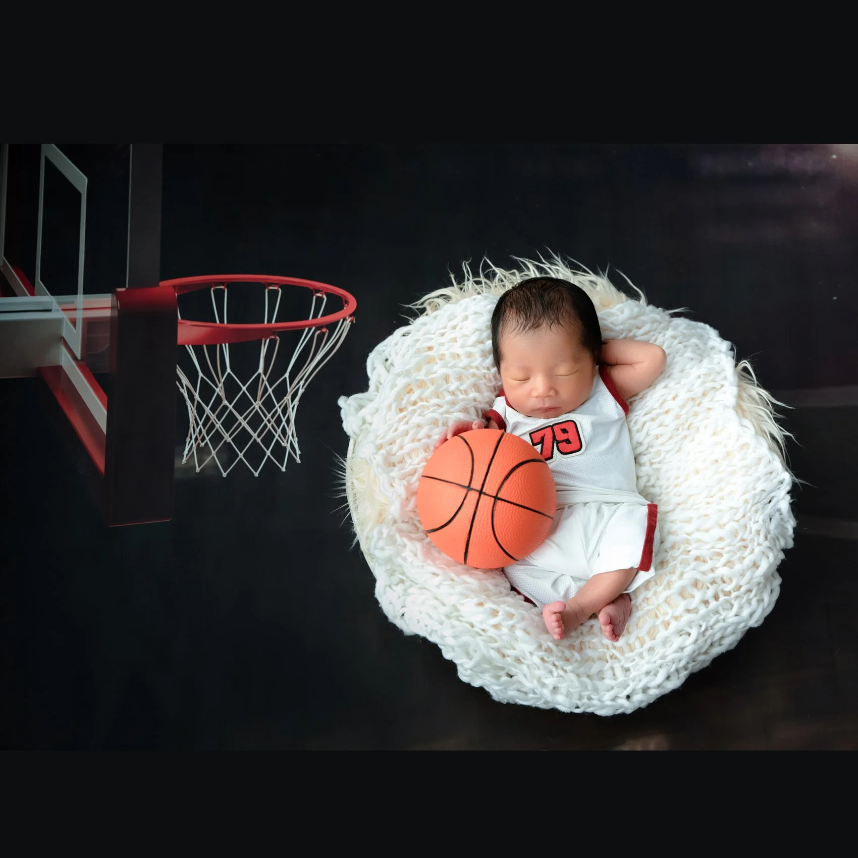 Sleeping newborn in a white basketball outfit reclining with one hand behind the head in a white basket beside an orange basketball and toy hoop