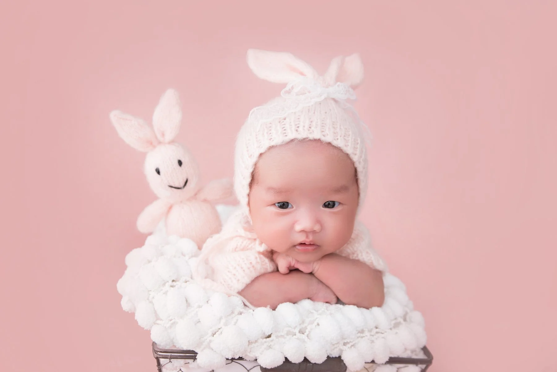 Awake newborn wearing a pale pink knitted bunny hat resting on folded arms in a white-lined basket beside a rabbit toy