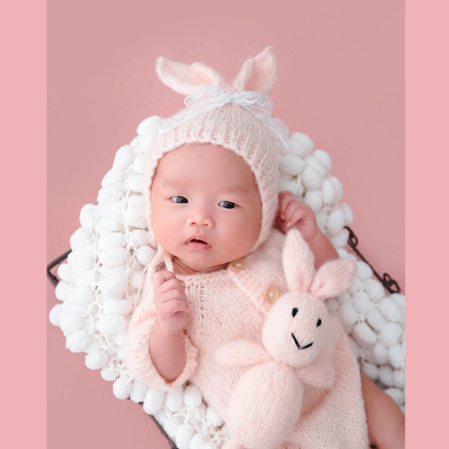 Awake newborn in a pale pink bunny hat and knitted outfit lying in a fluffy white basket with a rabbit toy