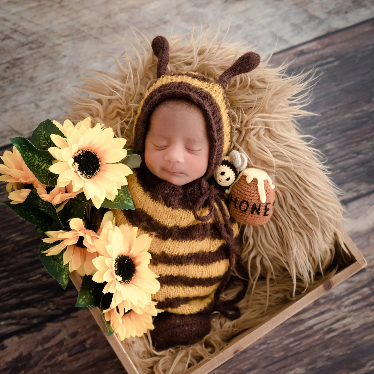 Sleeping newborn in a brown-and-yellow striped bee outfit in a rustic basket beside sunflowers and a honey pot