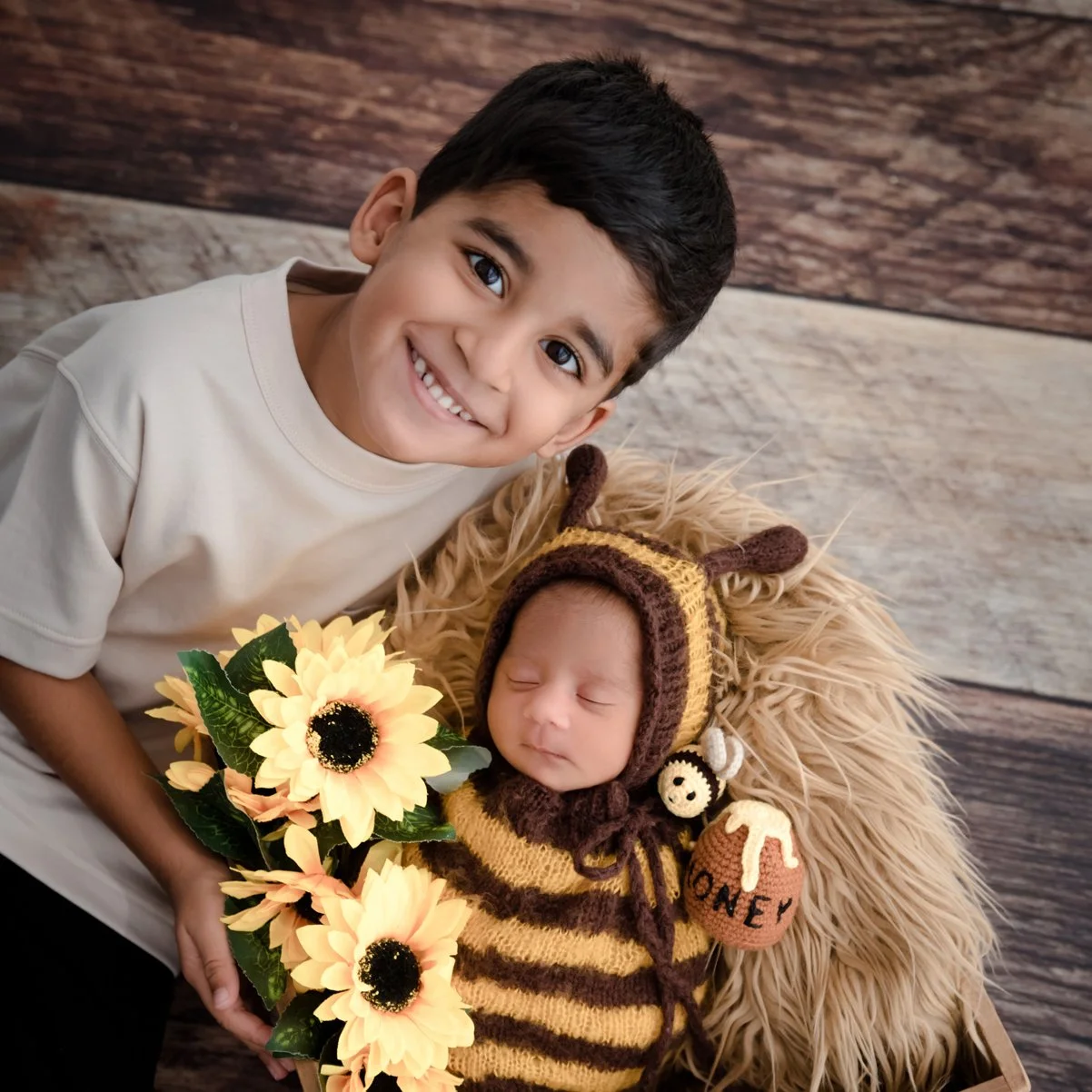 Smiling older sibling beside a sleeping newborn in a striped bee outfit nestled in a basket with sunflowers