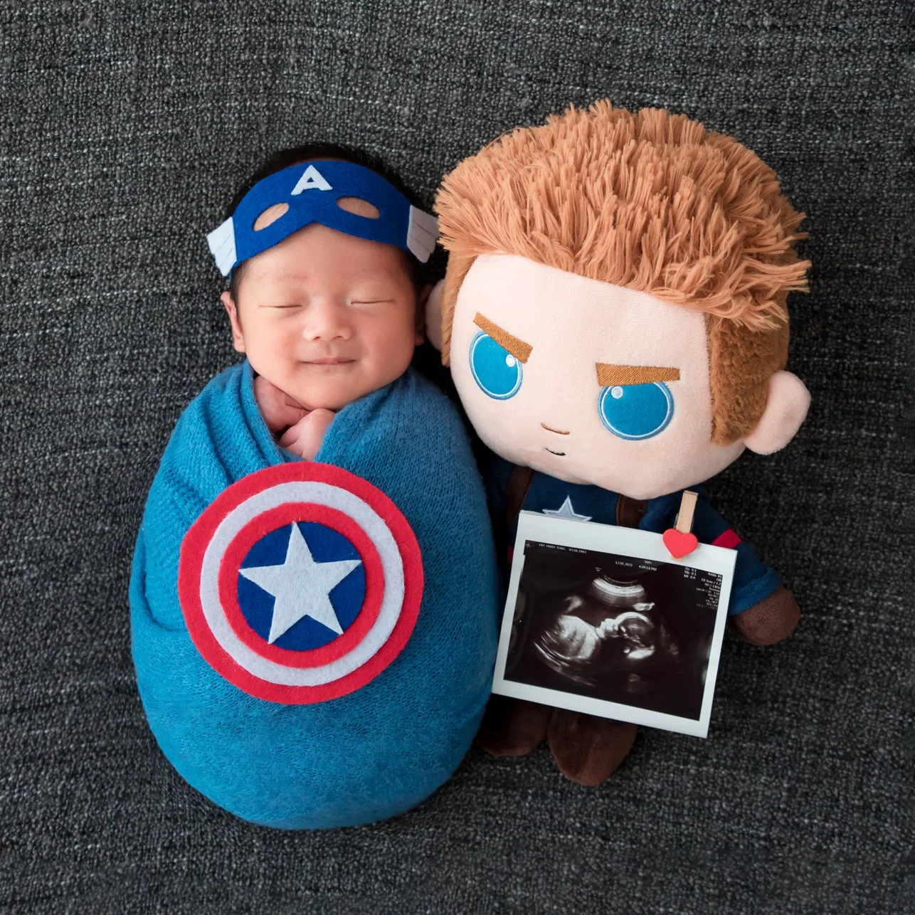 Blue-swaddled newborn wearing a Captain America hat with a shield beside a large superhero plush toy