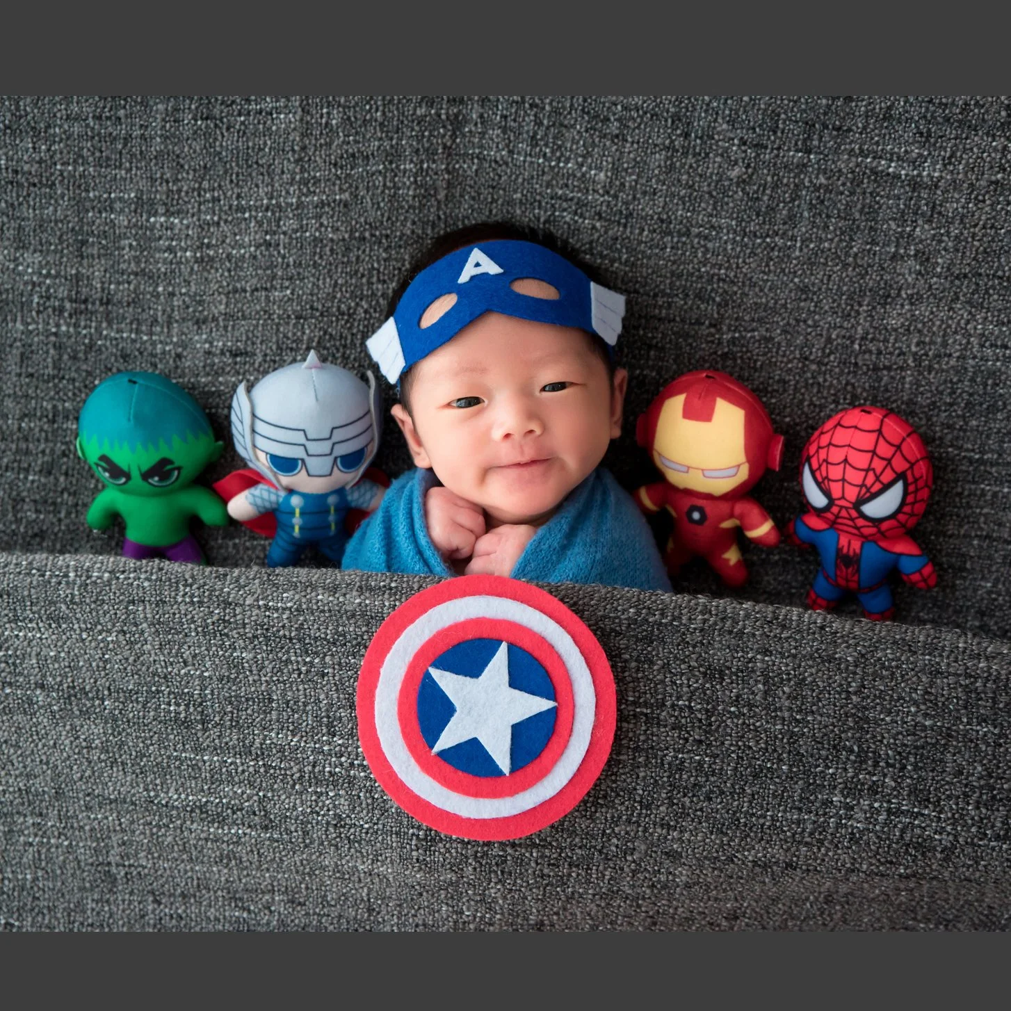 Awake newborn wearing a Captain America hat and shield surrounded by superhero plush toys on a grey backdrop