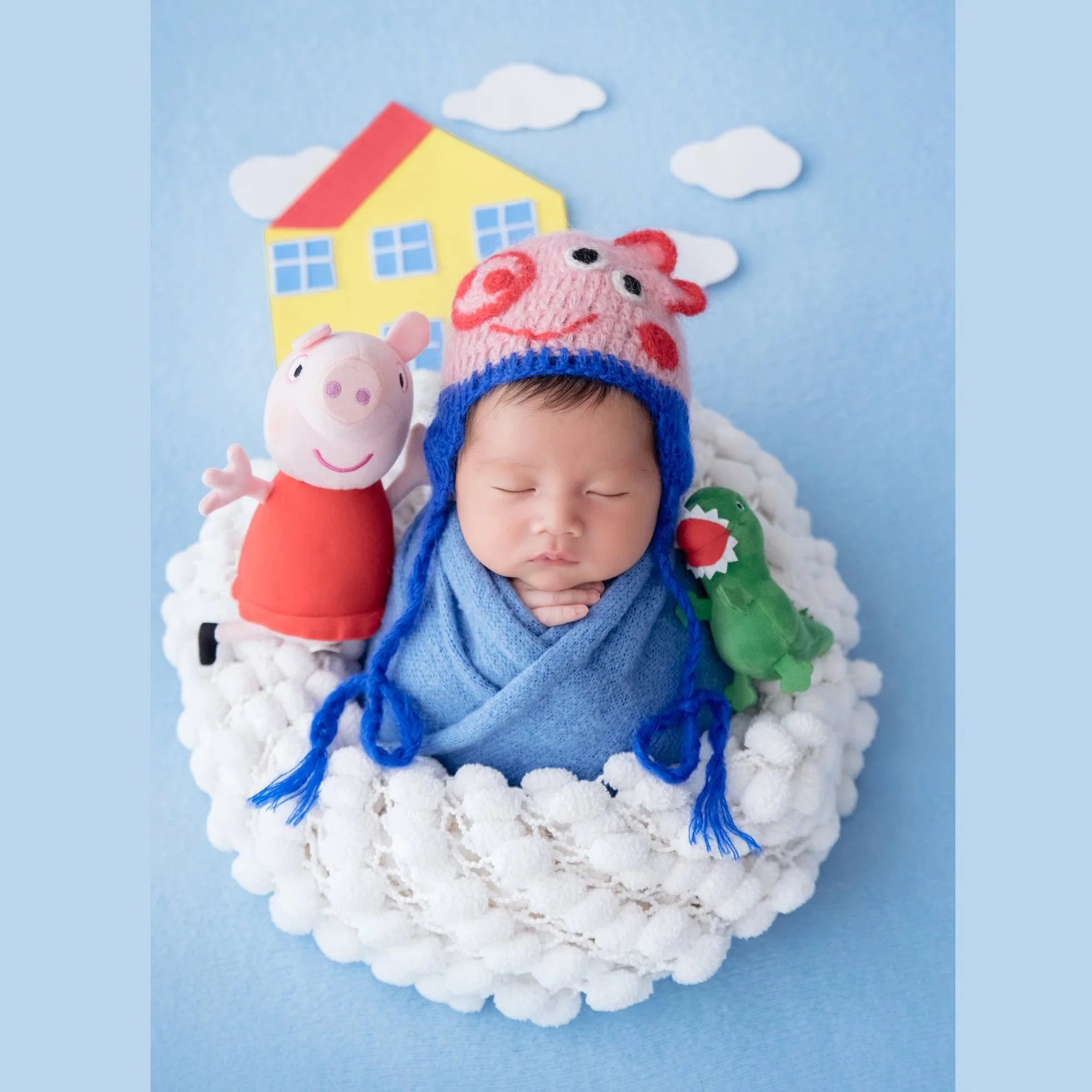 Blue-swaddled newborn wearing a George Pig hat in a white basket beside toy pigs and a dinosaur