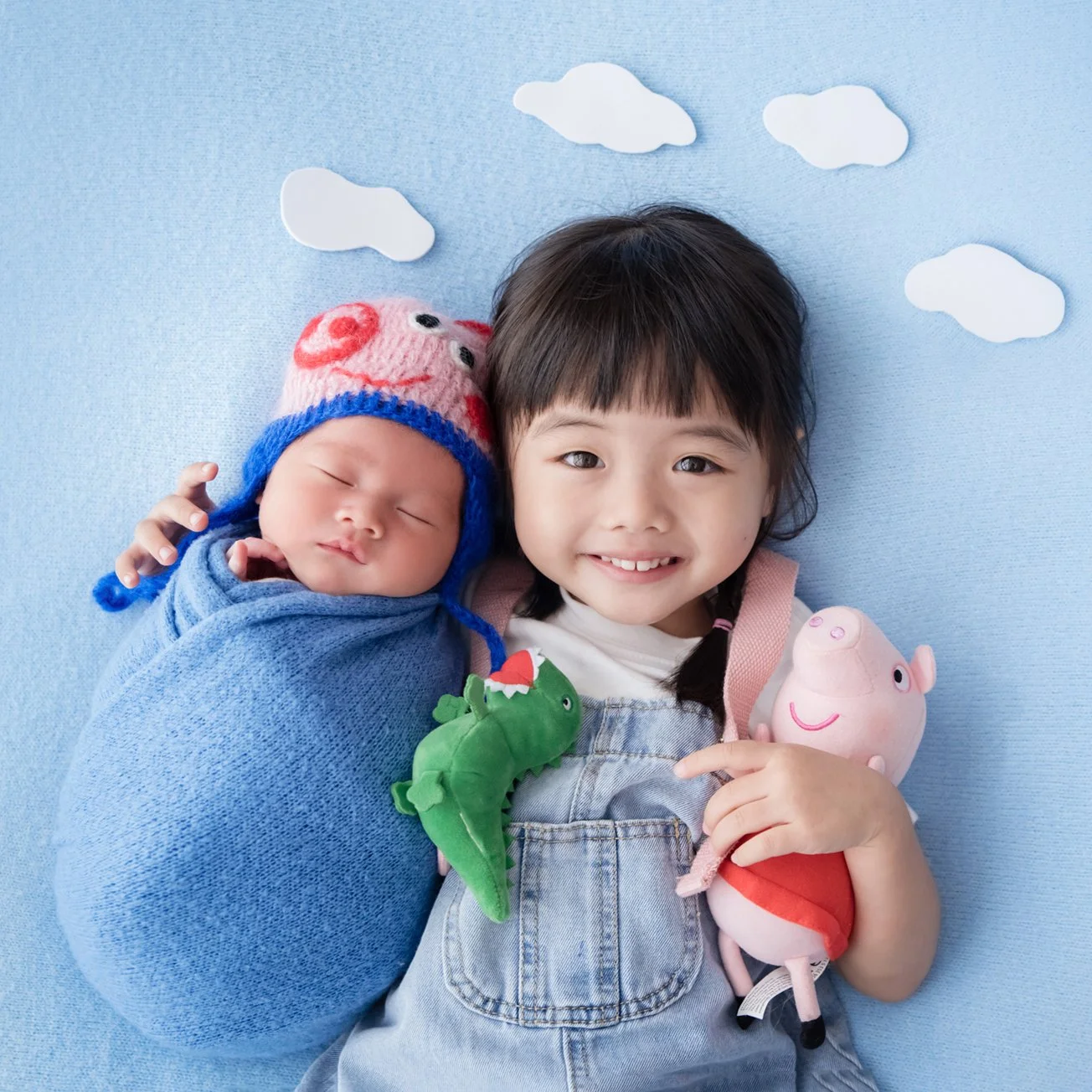 Smiling older sibling beside a blue-swaddled newborn in a George Pig hat, holding a toy pig and dinosaur