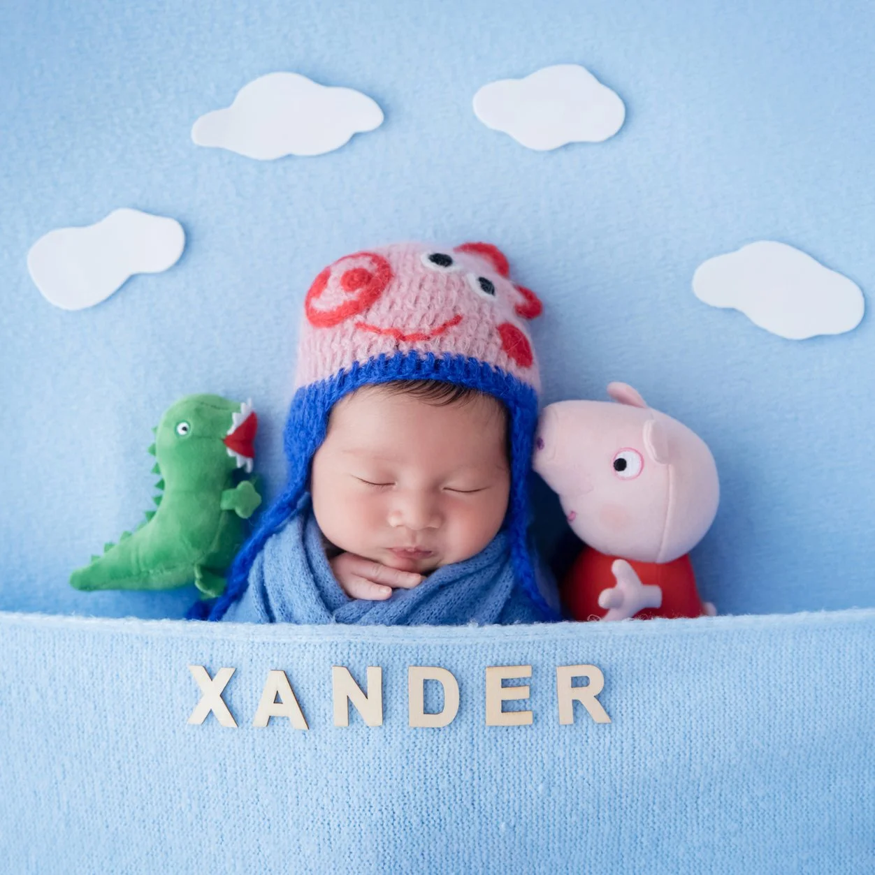 Sleeping newborn in a blue wrap and George Pig hat between toy pigs and a green dinosaur against a cloudy blue backdrop