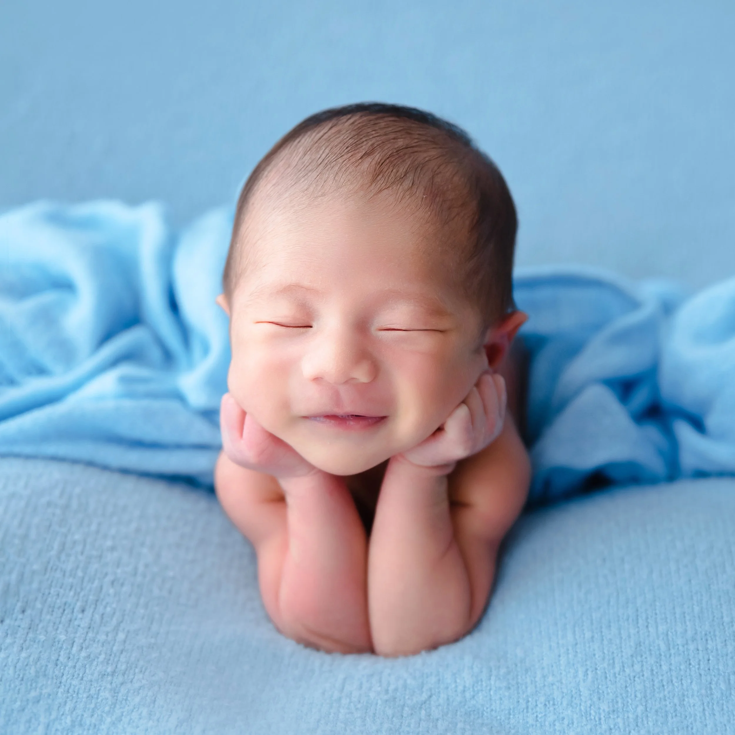 Smiling sleeping newborn resting both cheeks on hands on a blue blanket with blue fabric behind