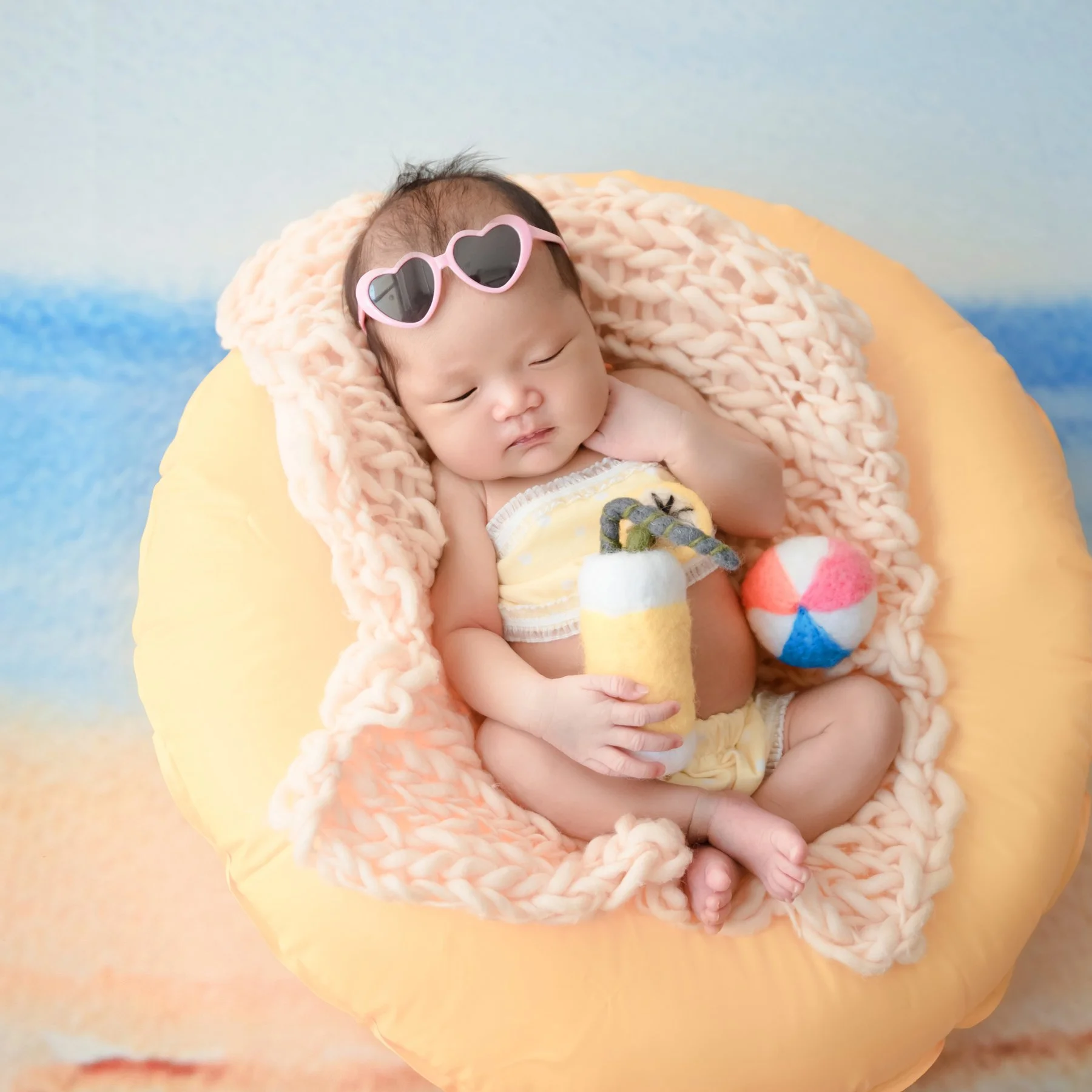Sleeping newborn with heart-shaped sunglasses on the head holding a toy drink in a yellow inflatable ring beside a miniature beach ball