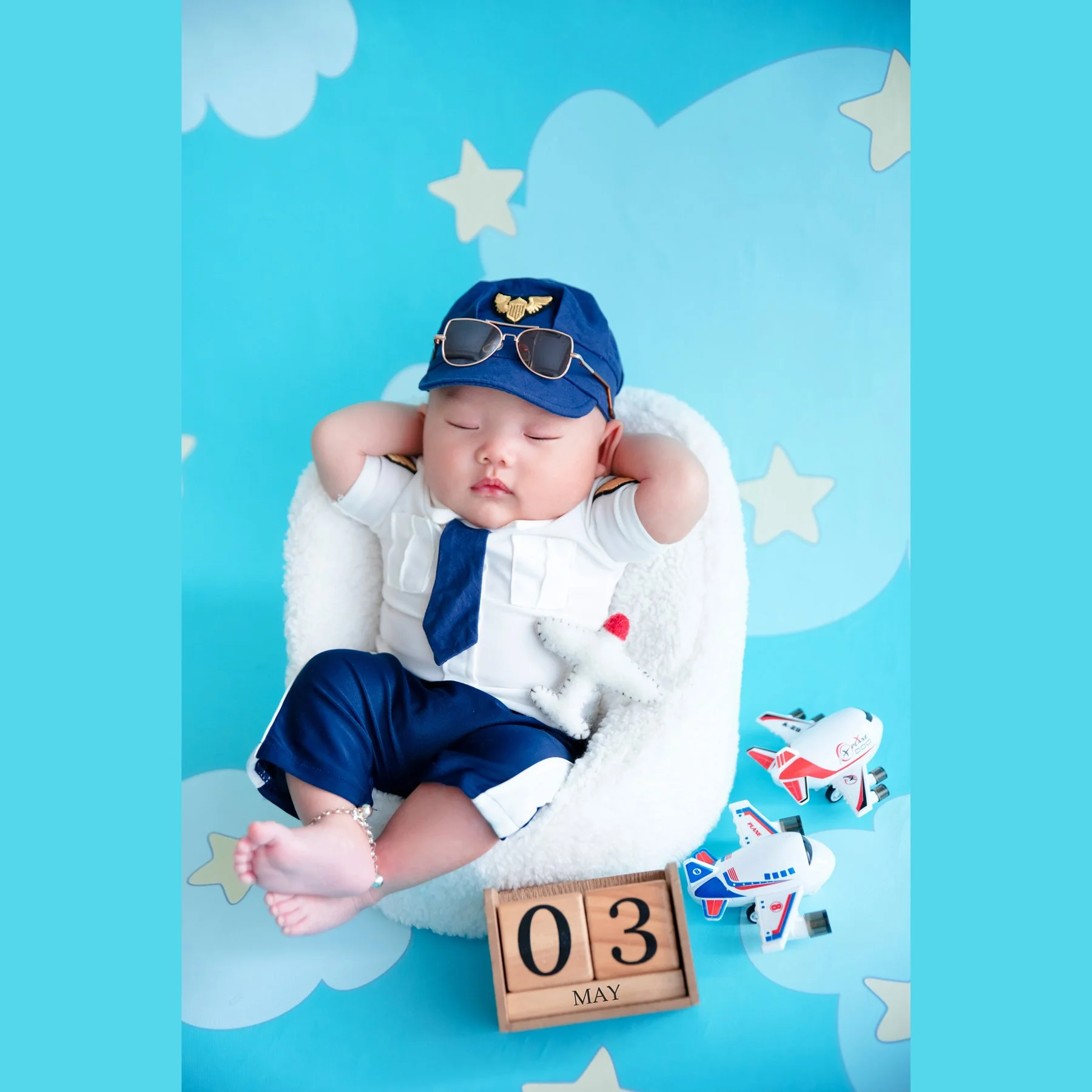 Sleeping newborn in a navy pilot cap, sunglasses and white uniform resting both hands behind the head in a basket on a blue cloud-and-star backdrop