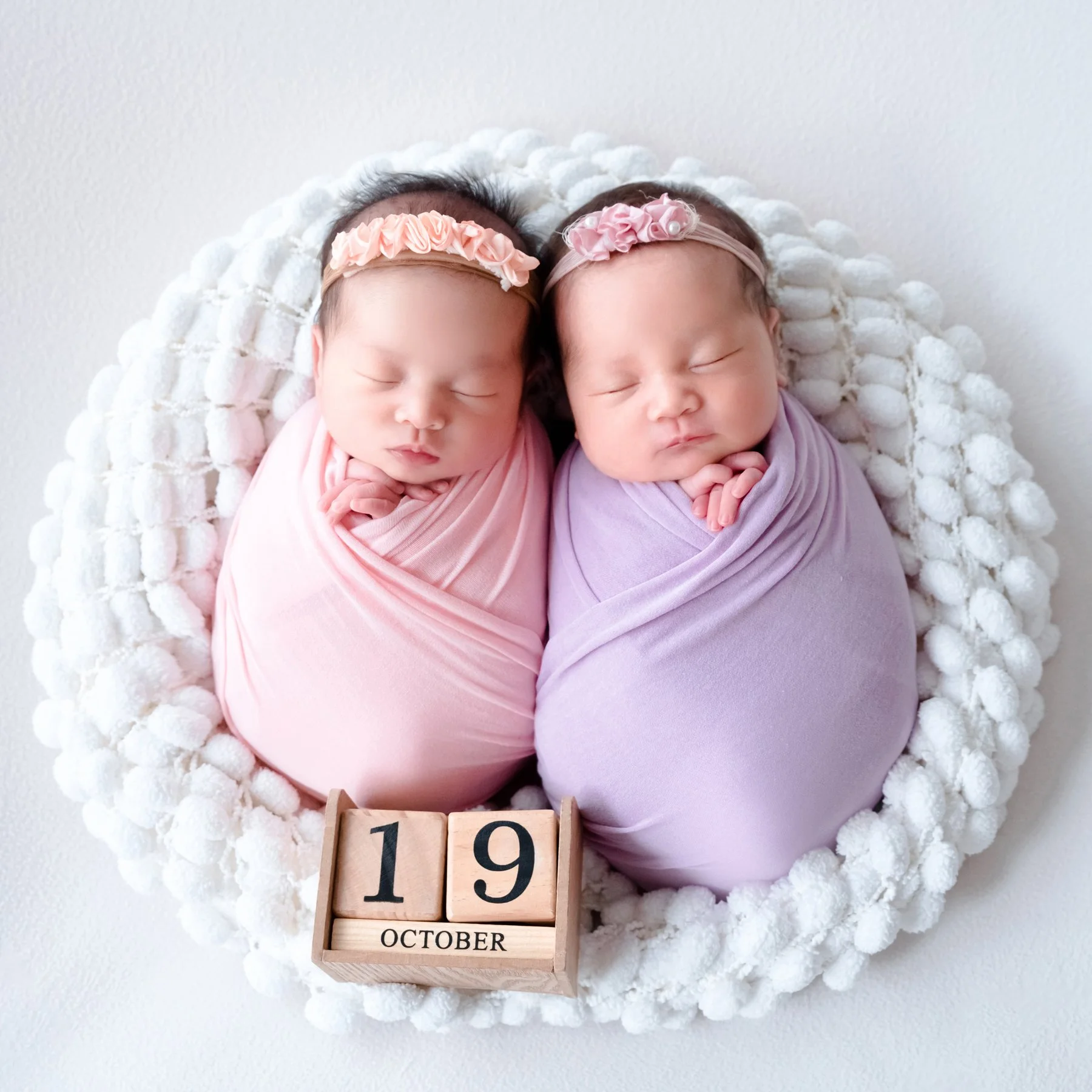 Newborn twins wrapped in pink and lavender blankets in a round white basket