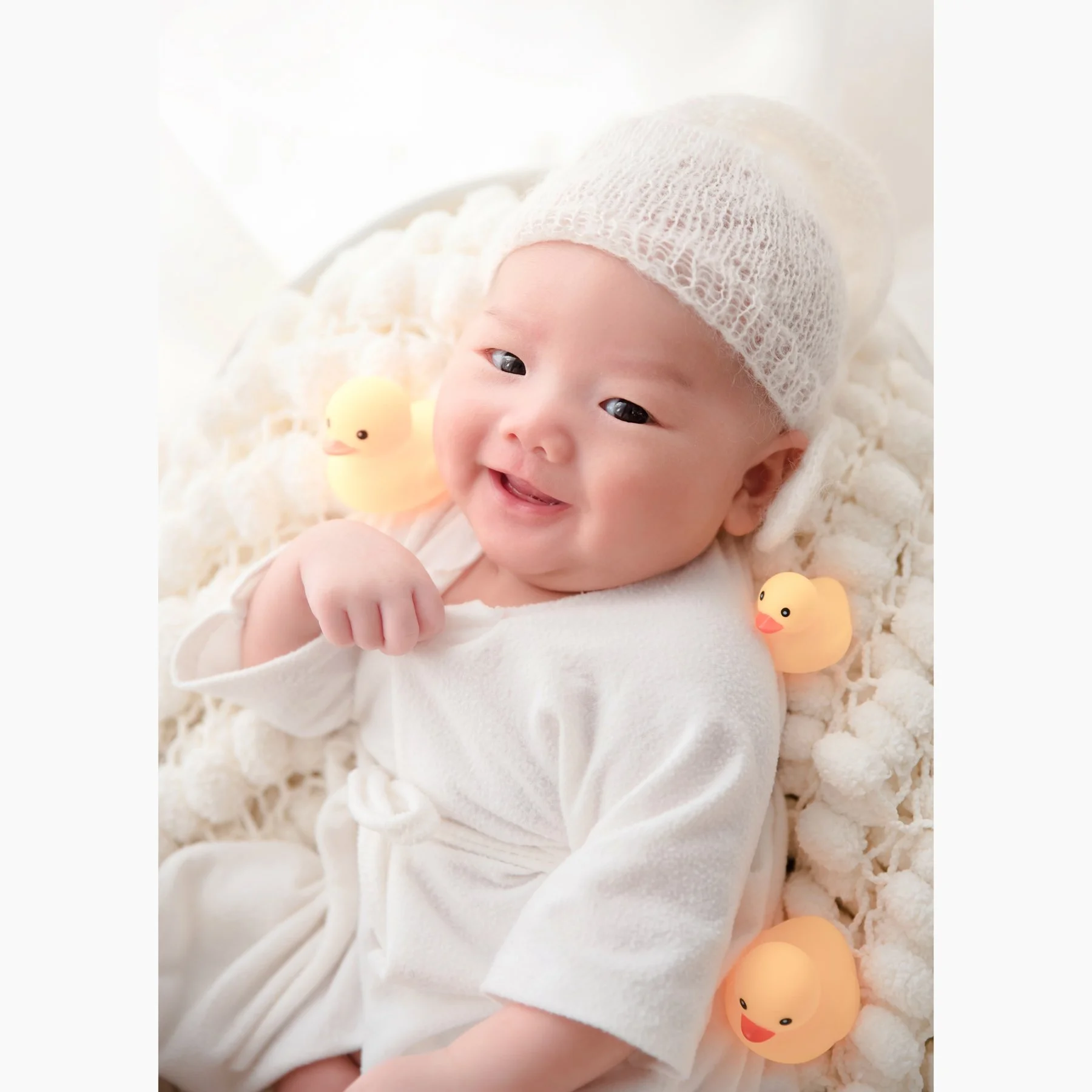 Smiling newborn in a white bathrobe and knitted hat reclining among white bath props and yellow rubber ducks