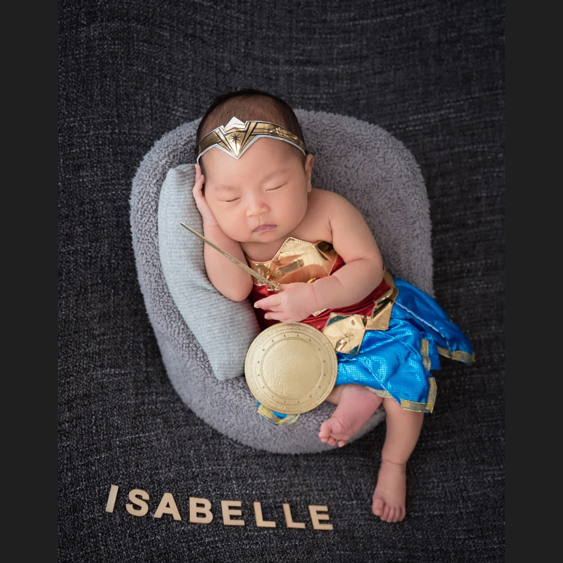 Sleeping newborn in a Wonder Woman costume with a gold headband and blue skirt in a grey basket