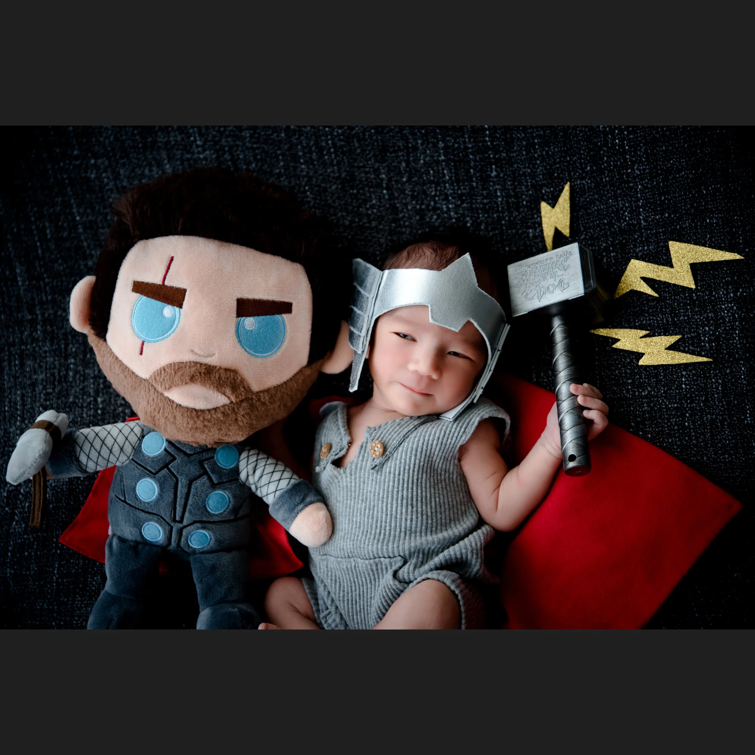 Newborn in a grey Thor outfit and helmet holding a toy hammer beside a large Thor plush and red cape