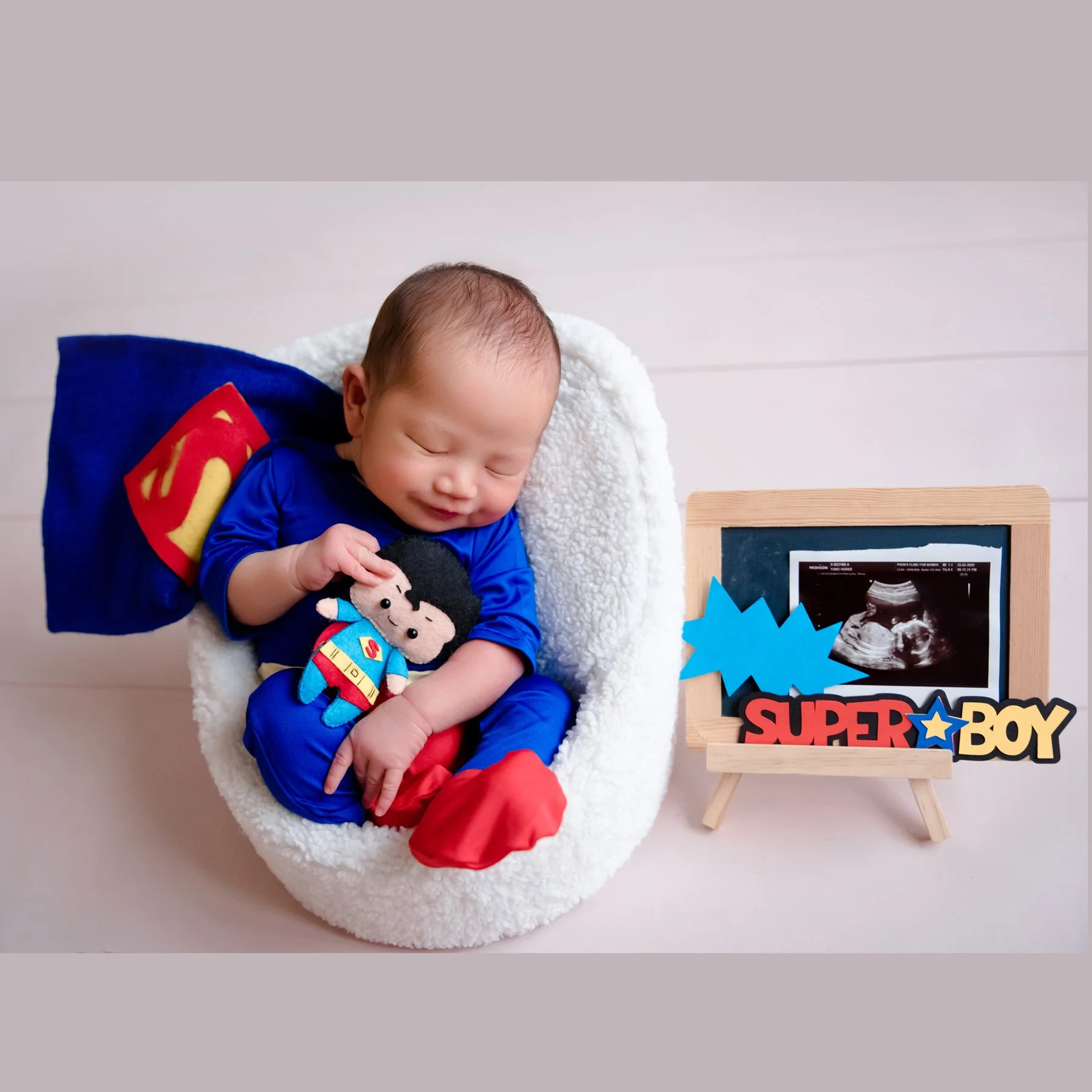 Sleeping newborn in a blue Superman outfit and red socks in a white basket beside a superhero cushion