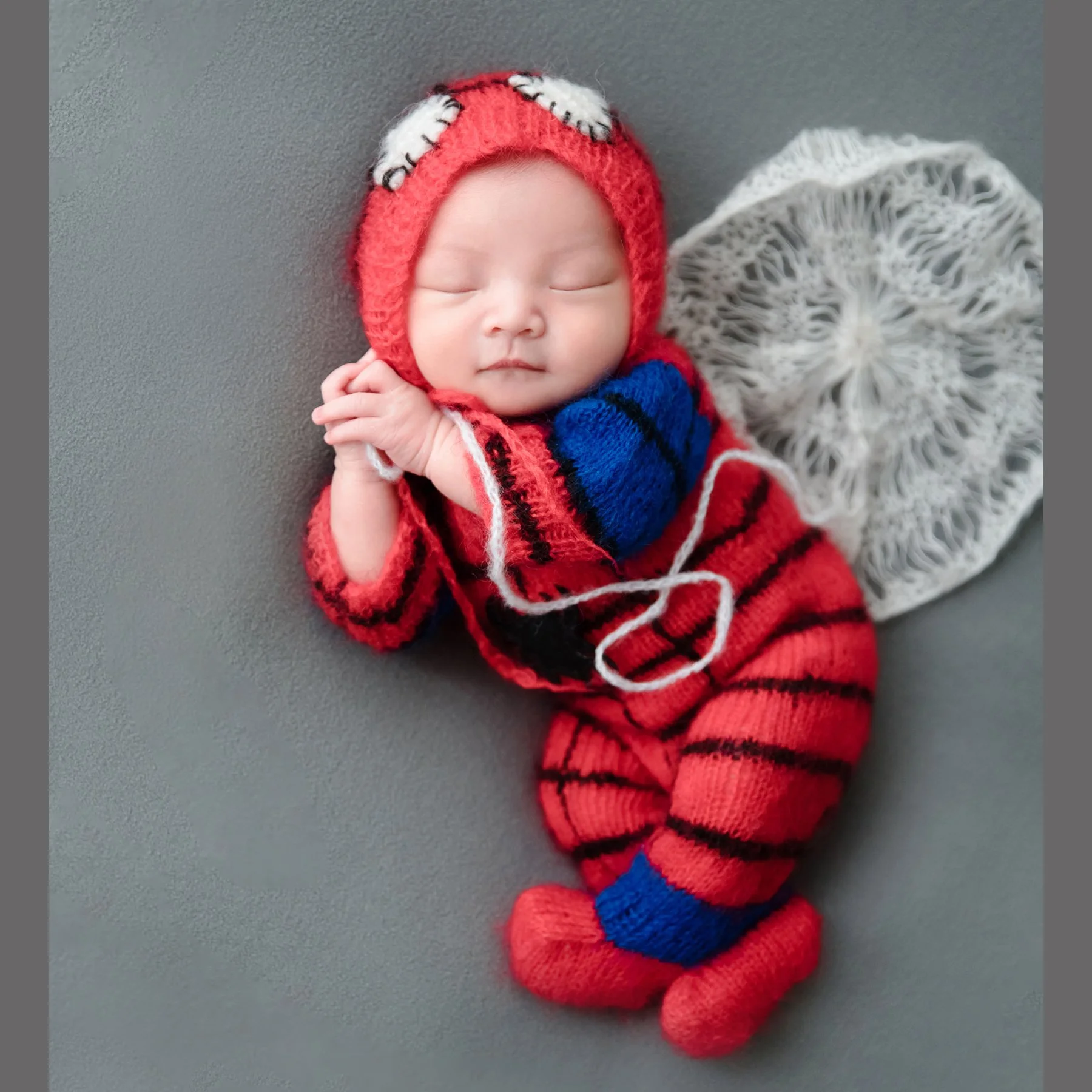 Sleeping newborn in a red-and-blue knitted Spider-Man costume holding a string beside a white web prop