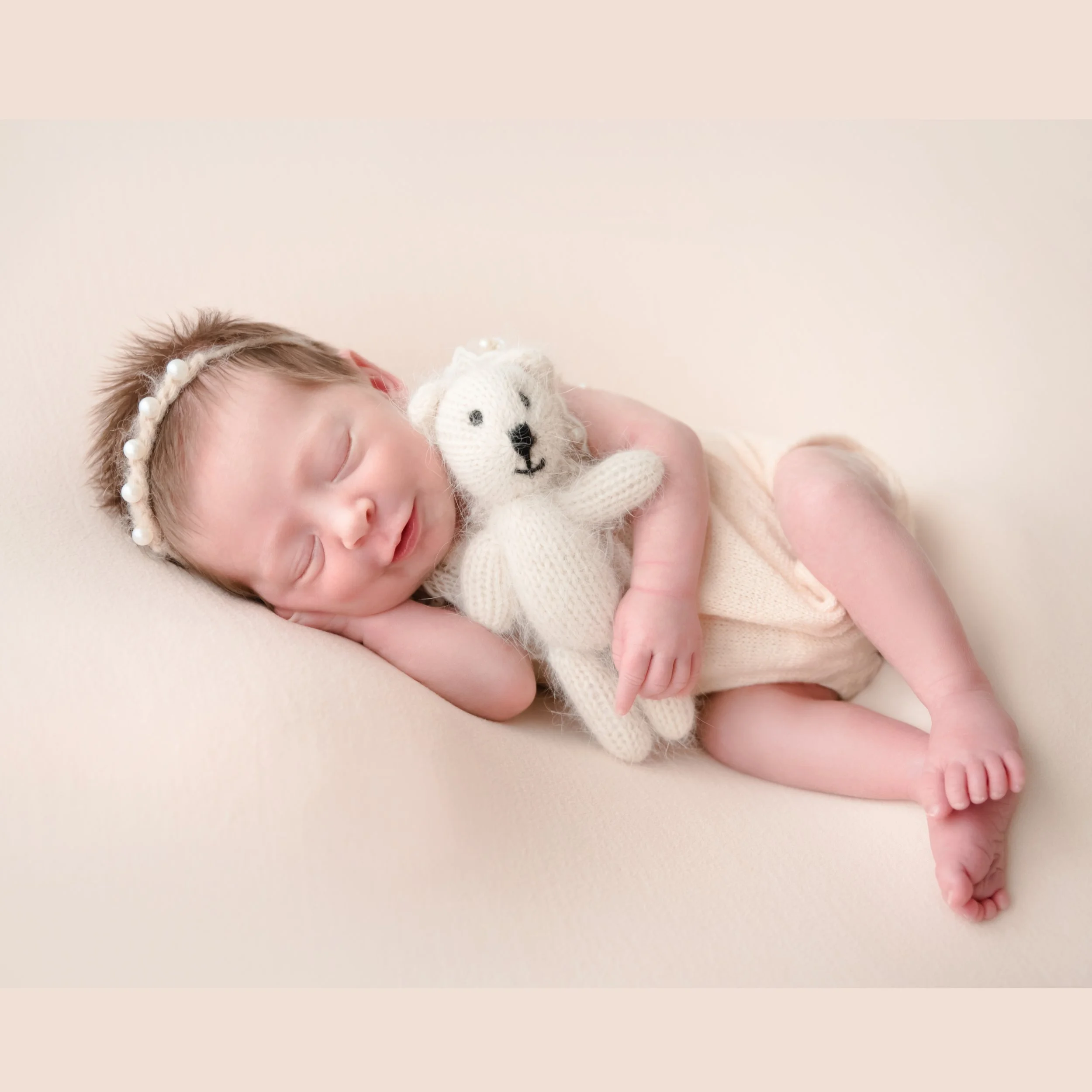 Sleeping newborn in a cream knitted outfit and pearl headband curled on one side while cuddling a small cream teddy bear on a beige backdrop
