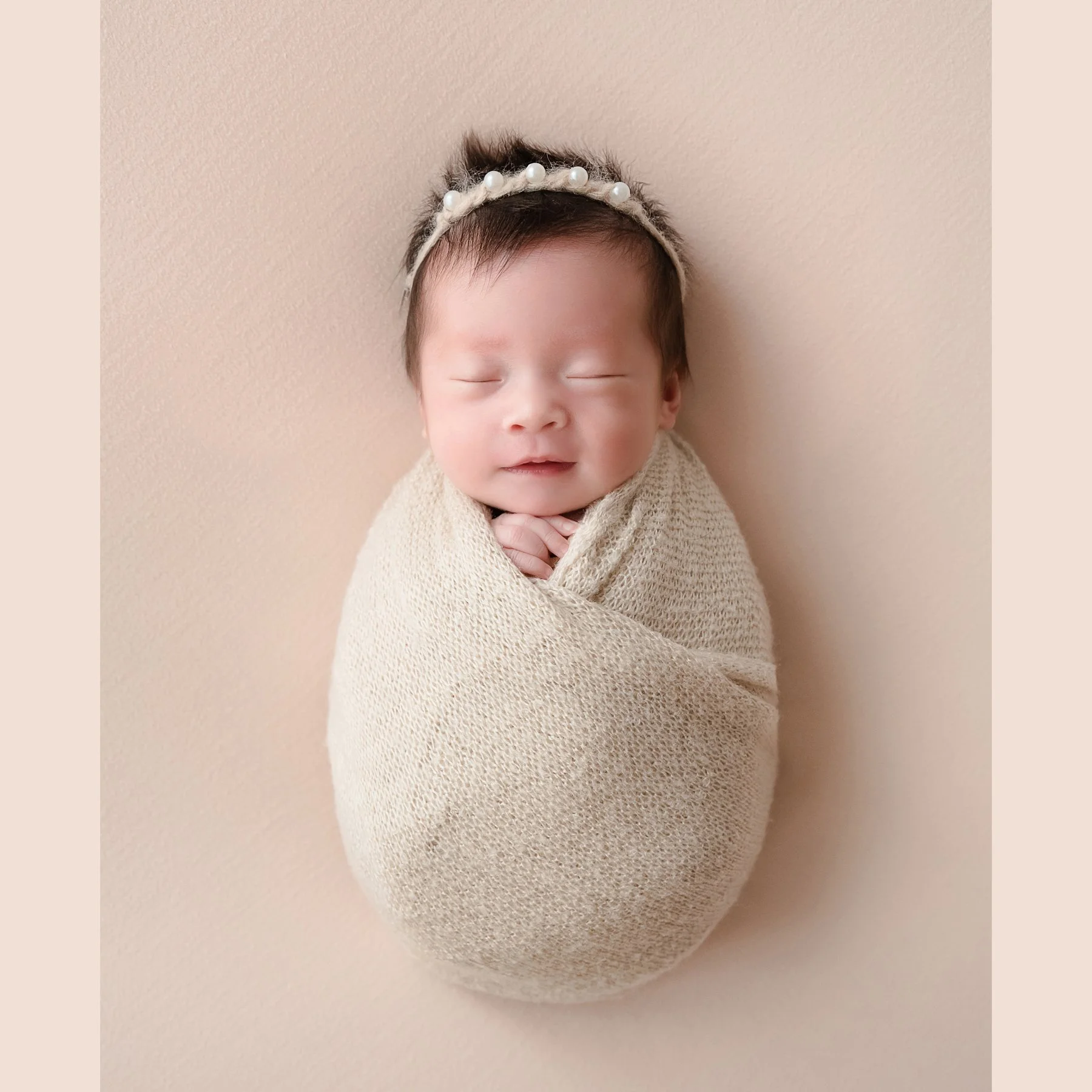 Smiling sleeping newborn in a cream swaddle and pearl headband reclining on a plain beige backdrop