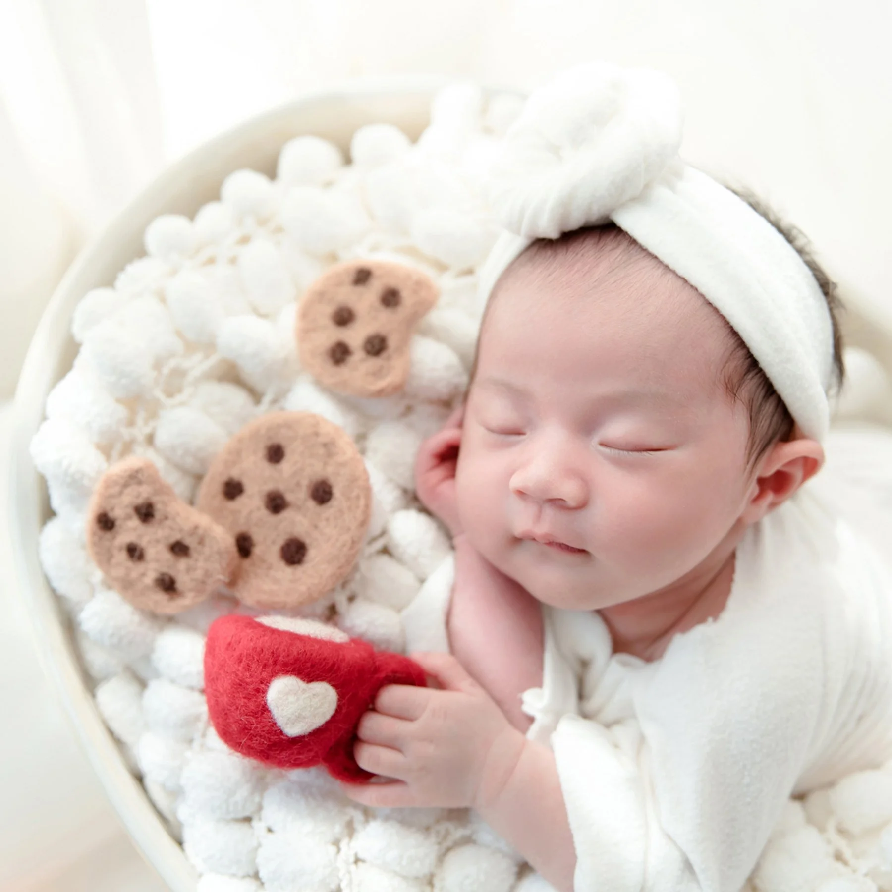 Sleeping newborn in a white bathrobe and bow headband cuddling a red cup among white bath props and toy cookies