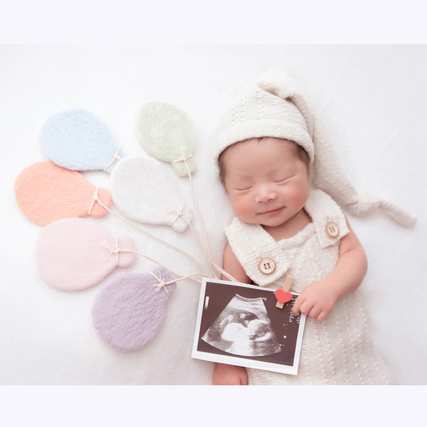 Smiling sleeping newborn in a white knitted hat and overalls beside an ultrasound picture and pastel balloon decorations on a white backdrop
