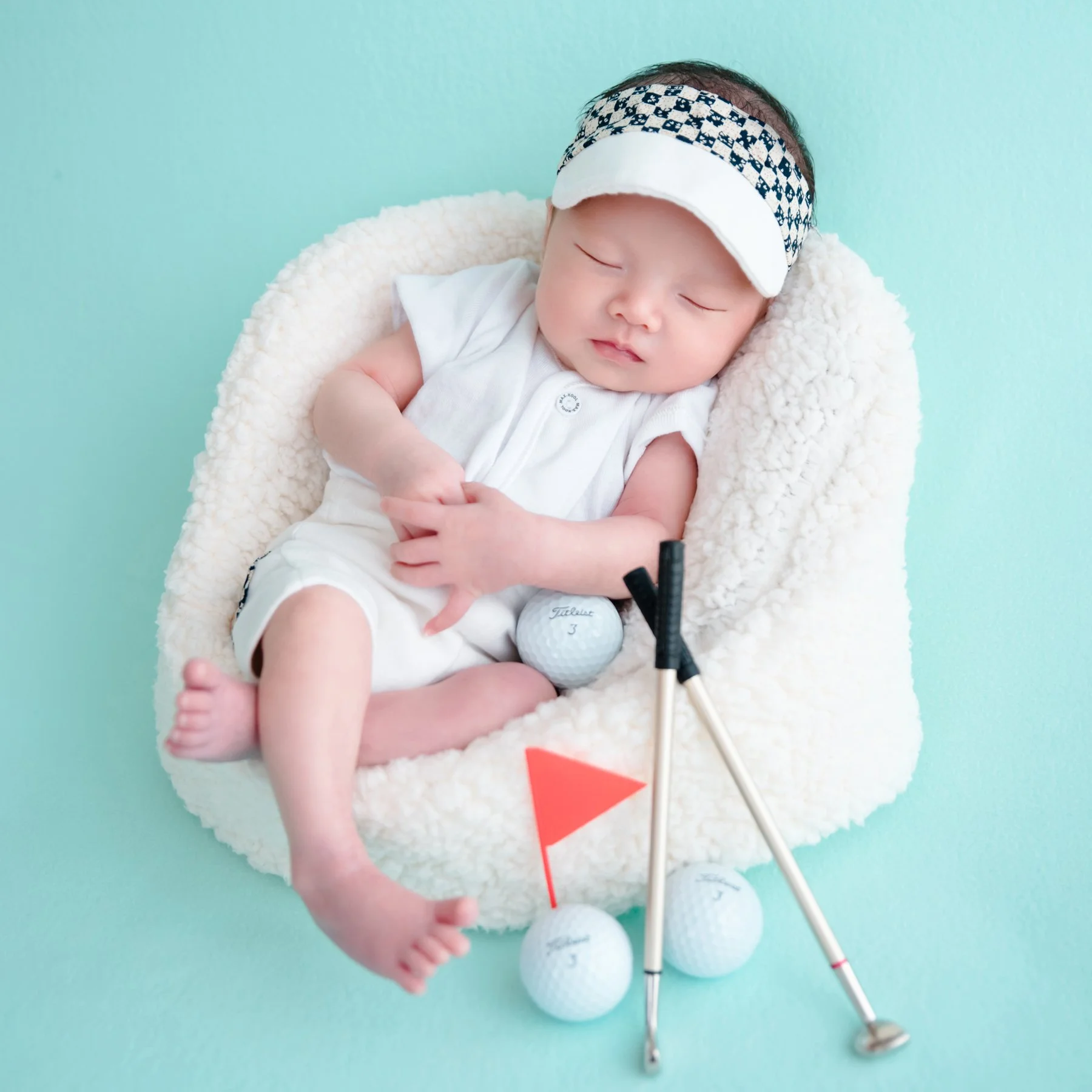 Sleeping newborn in a white golf outfit and checked visor reclining in a white basket beside miniature golf clubs, balls and a red flag on a turquoise backdrop
