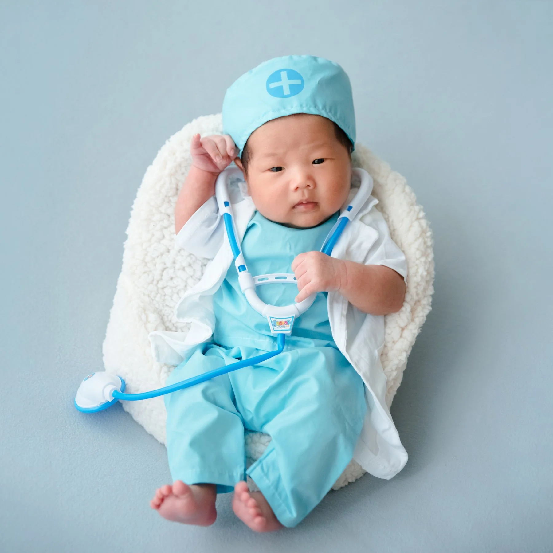 Awake newborn in blue medical scrubs, a cap and white coat reclining in a fluffy white basket with a toy stethoscope