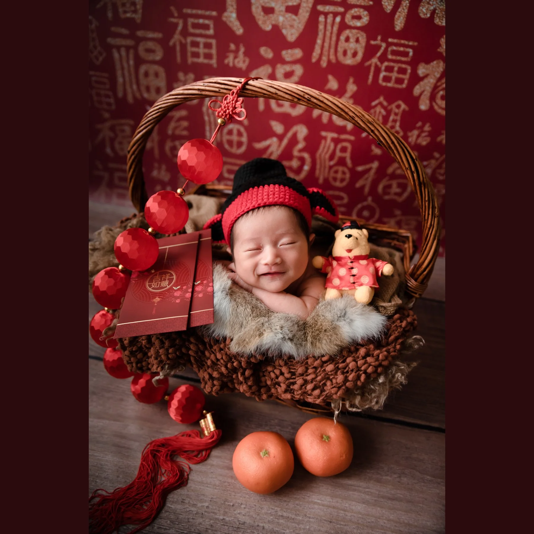Smiling newborn wearing a red knitted hat with chin on folded arms in a woven basket beside red lanterns, oranges and a festive teddy bear