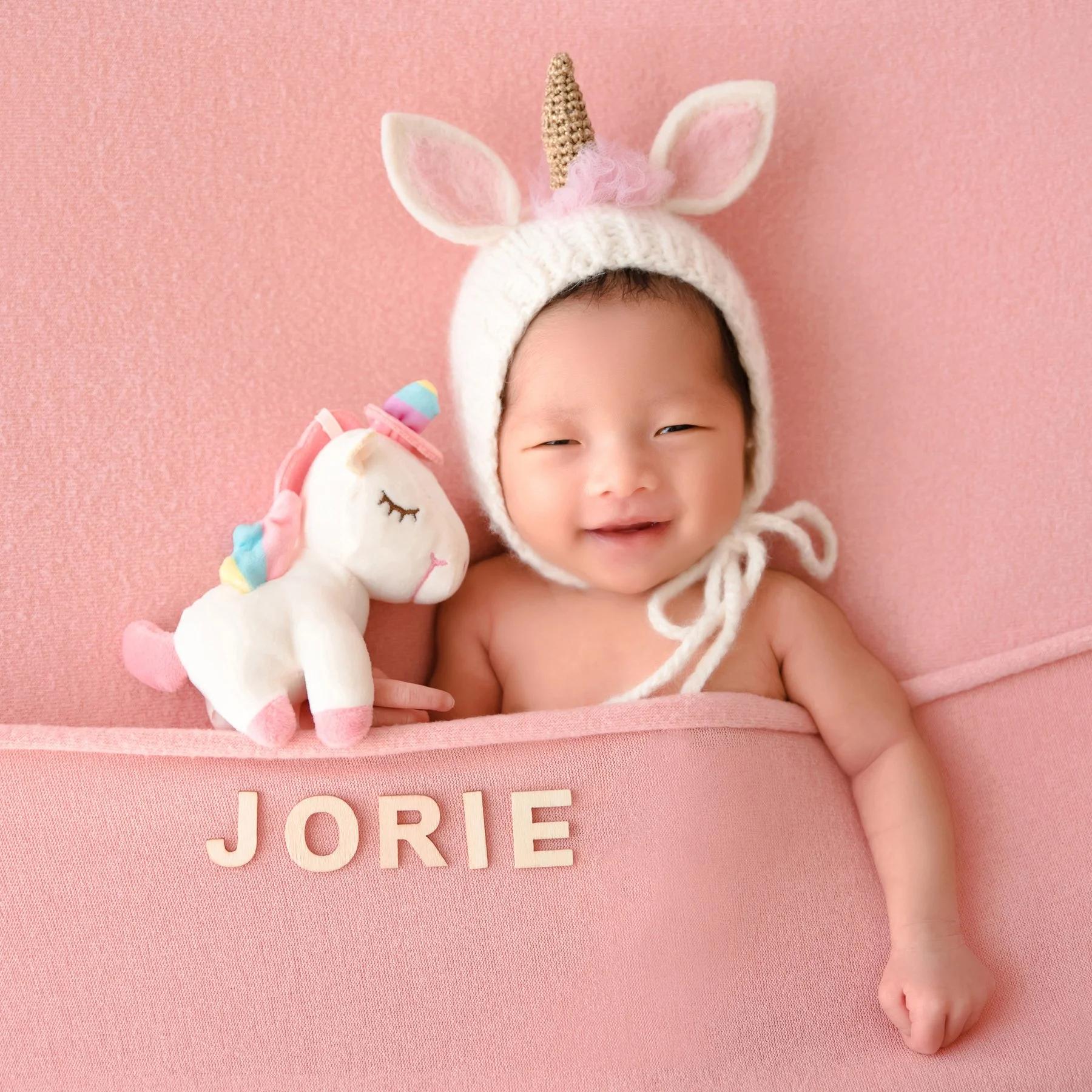 Smiling newborn in a white unicorn hat beside a unicorn plush against a pink backdrop