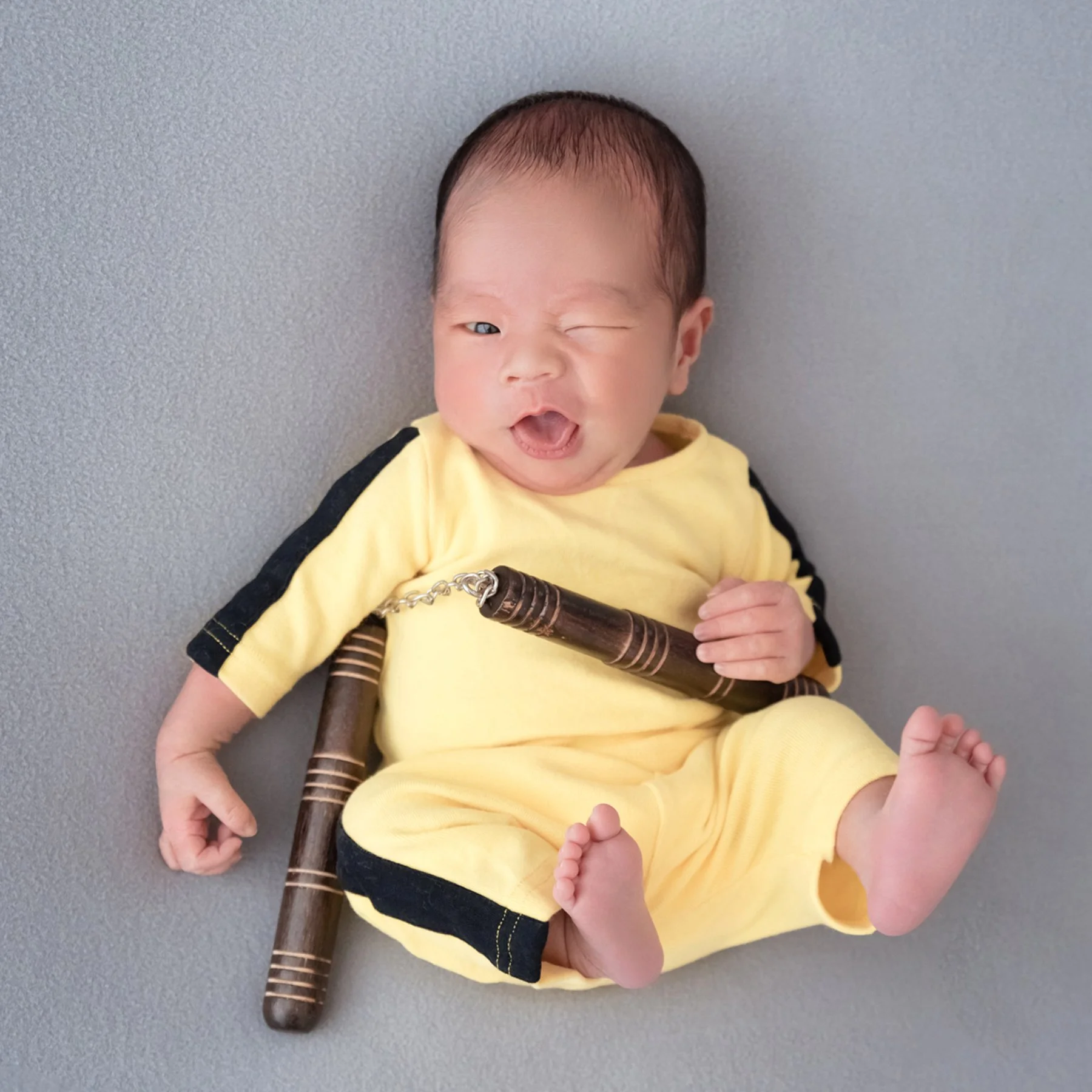 Awake newborn in a yellow-and-black Bruce Lee inspired martial arts outfit holding toy nunchucks on a grey backdrop