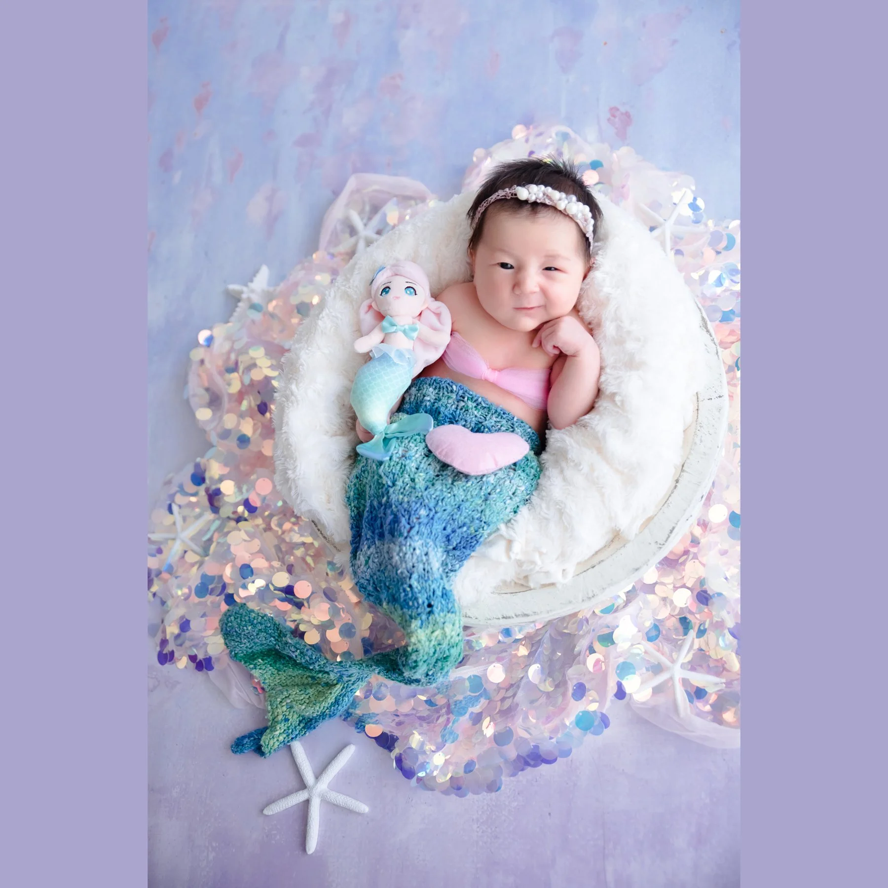 Awake newborn in a turquoise mermaid tail and purple top lying in a white basket surrounded by pastel flowers and starfish