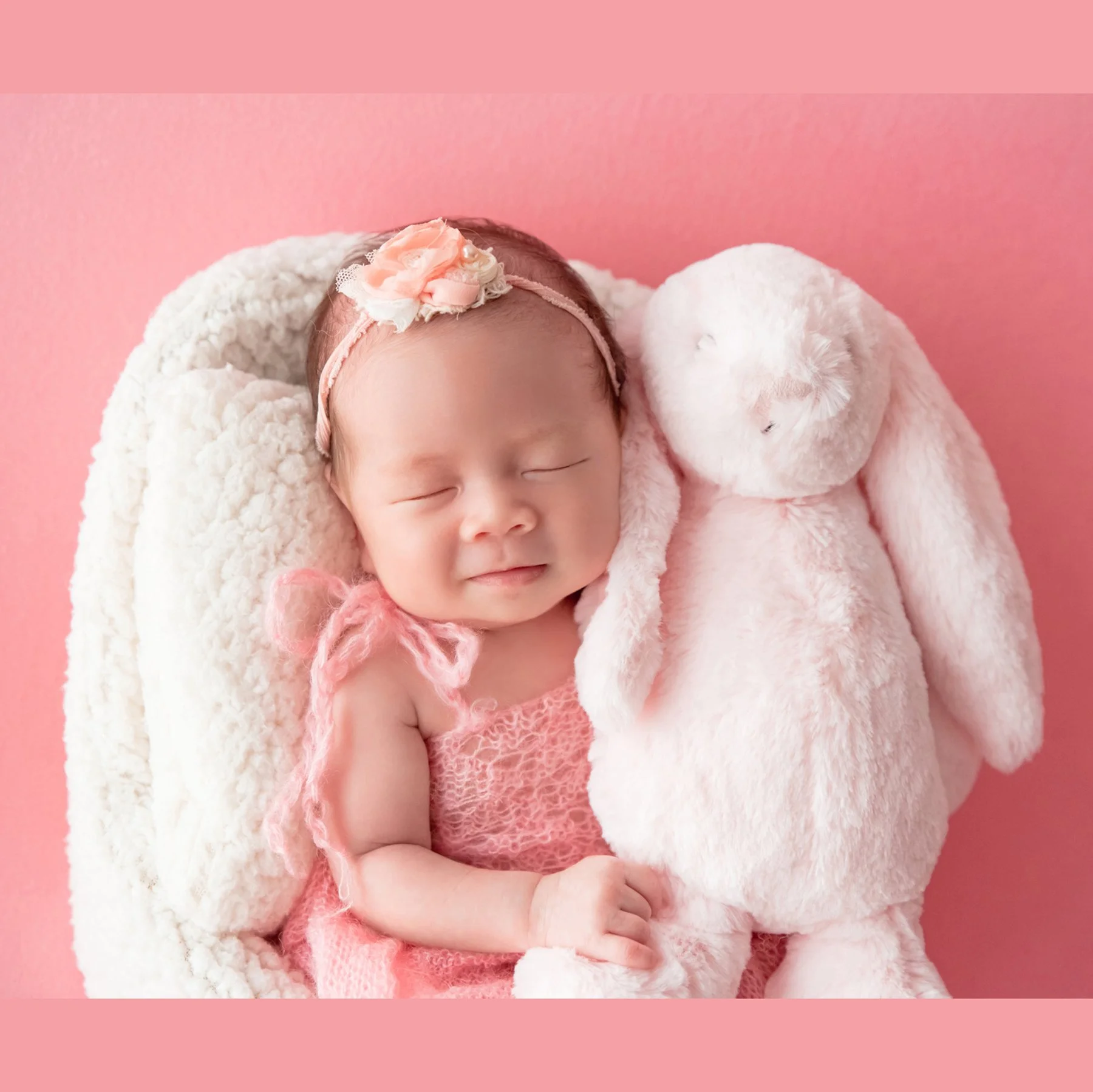 Sleeping newborn in a pink knitted outfit and peach floral headband reclining in a white basket while cuddling a pale pink rabbit plush