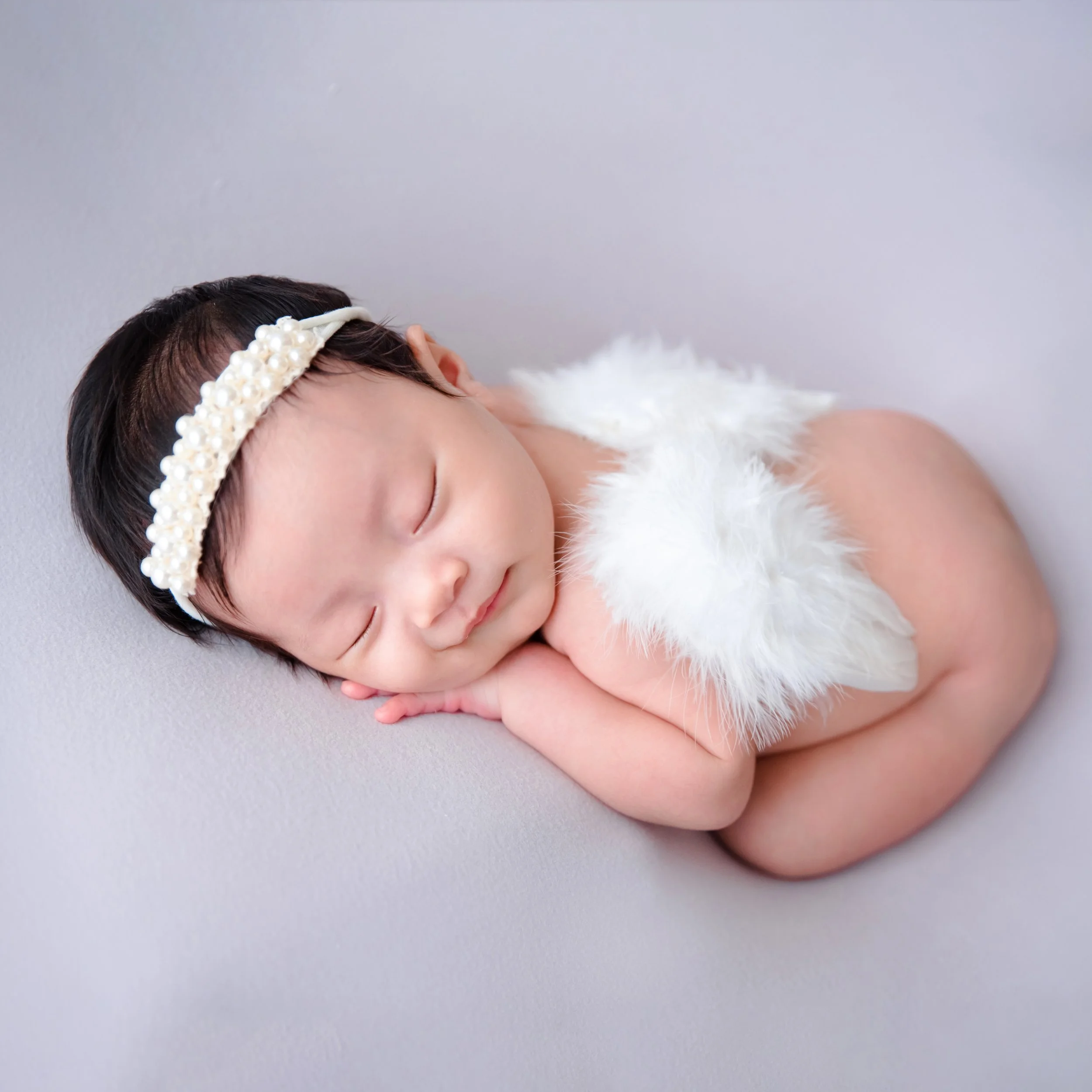 Sleeping newborn with white angel wings and a lace headband curled on one side against pale lavender