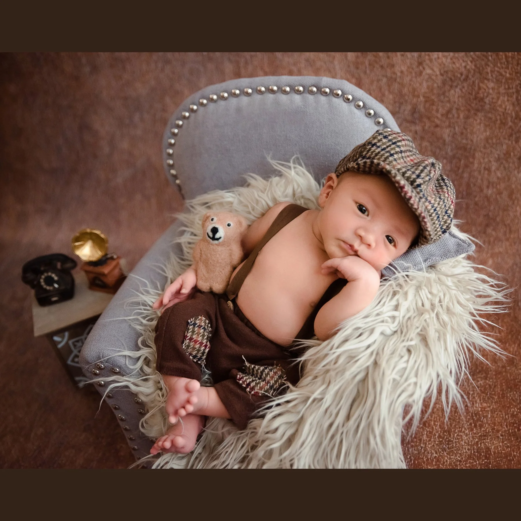 Awake newborn in a checked cap and brown wrap reclining in a miniature grey armchair with one hand beside the cheek, a teddy bear and vintage telephone nearby