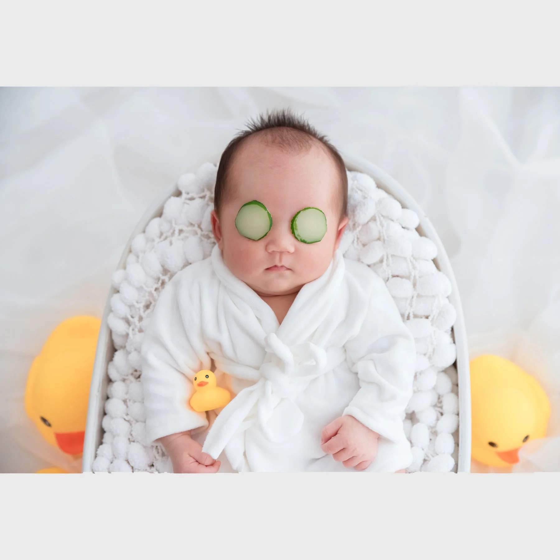 Newborn in a white bathrobe reclining in a miniature bathtub with cucumber-slice props over the eyes and yellow rubber ducks beside the tub