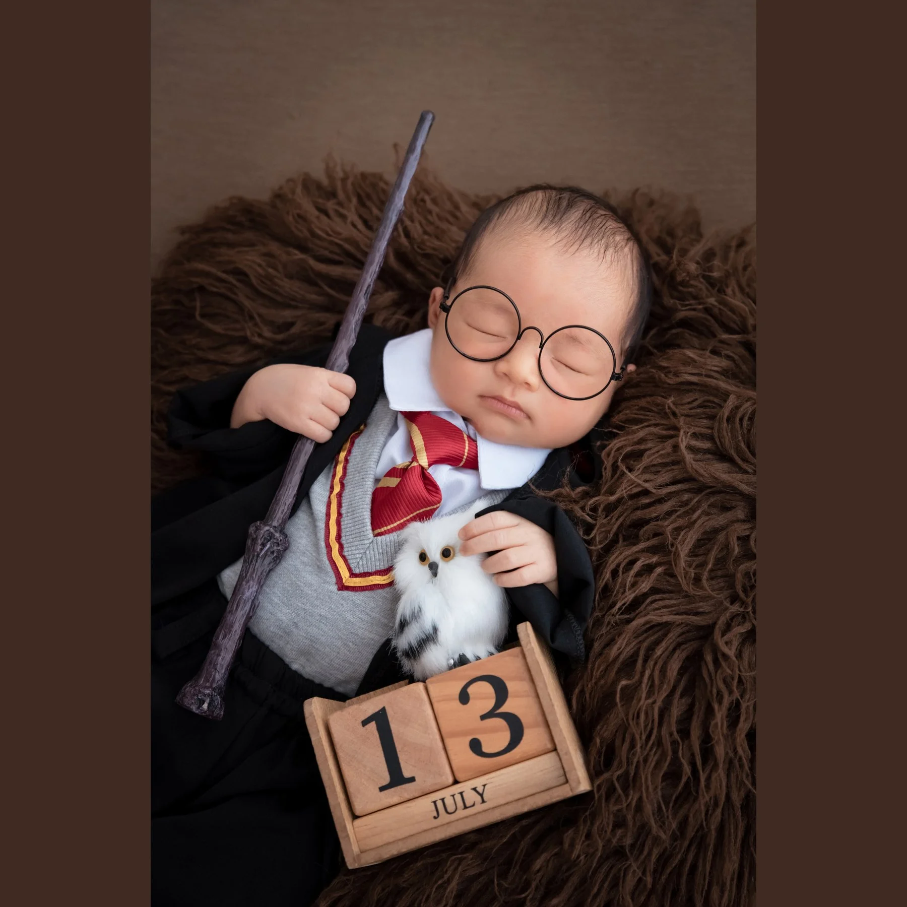 Sleeping newborn in a Harry Potter robe and round glasses holding a toy wand beside an owl on brown fur