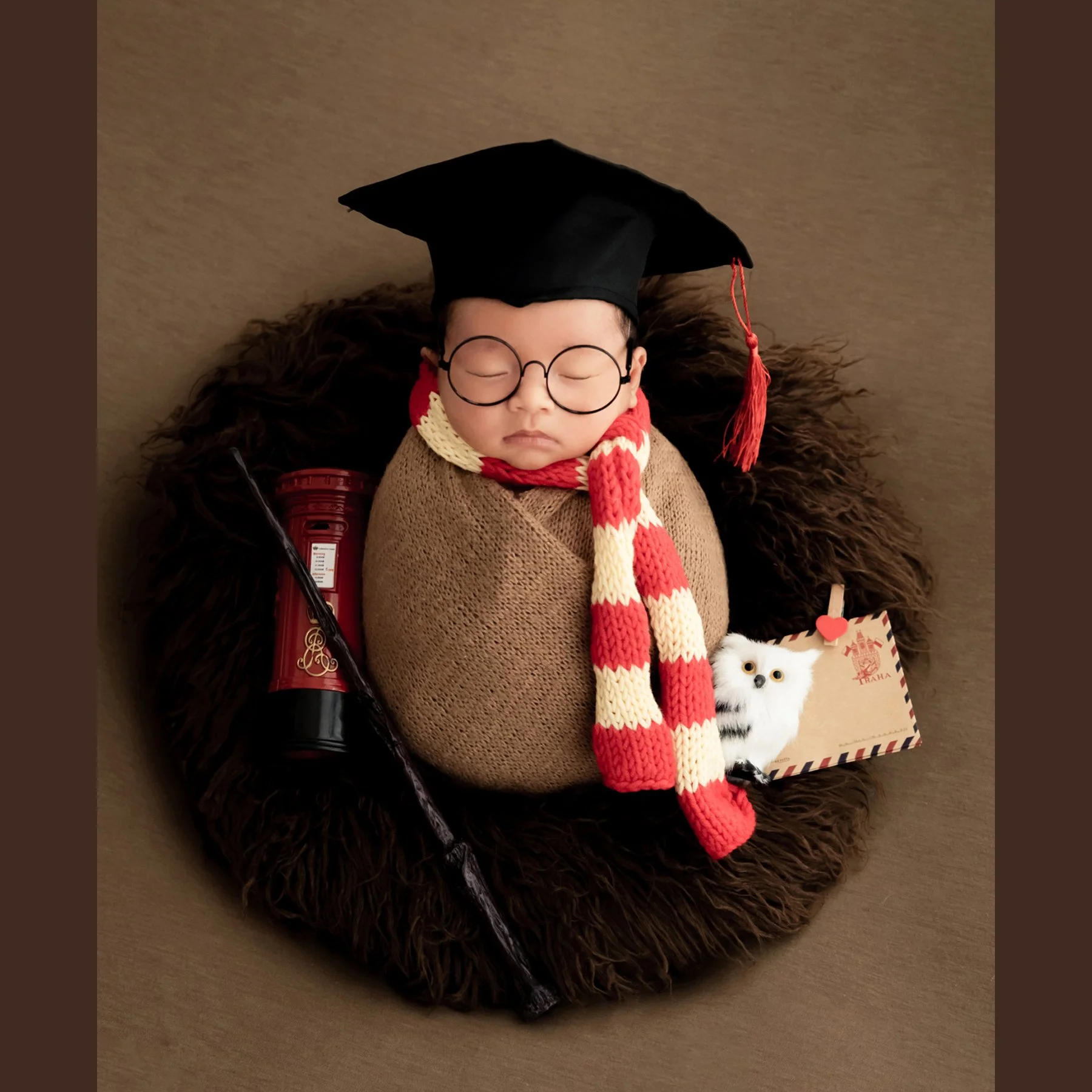 Newborn wrapped in brown fabric wearing round glasses, a graduation cap and striped scarf beside a toy wand